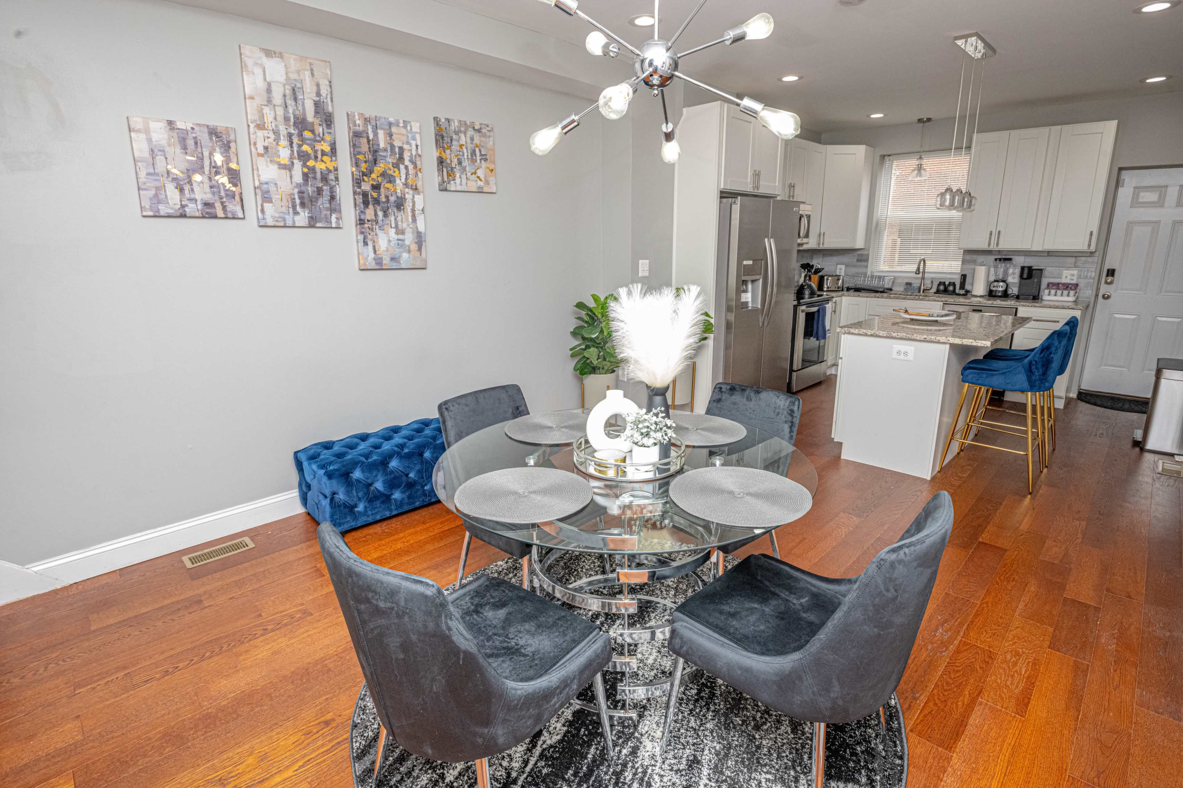 A modern dining area with a glass table surrounded by dark chairs, complemented by a stylish chandelier, sleek kitchen appliances visible in the background, and decorative wall art.