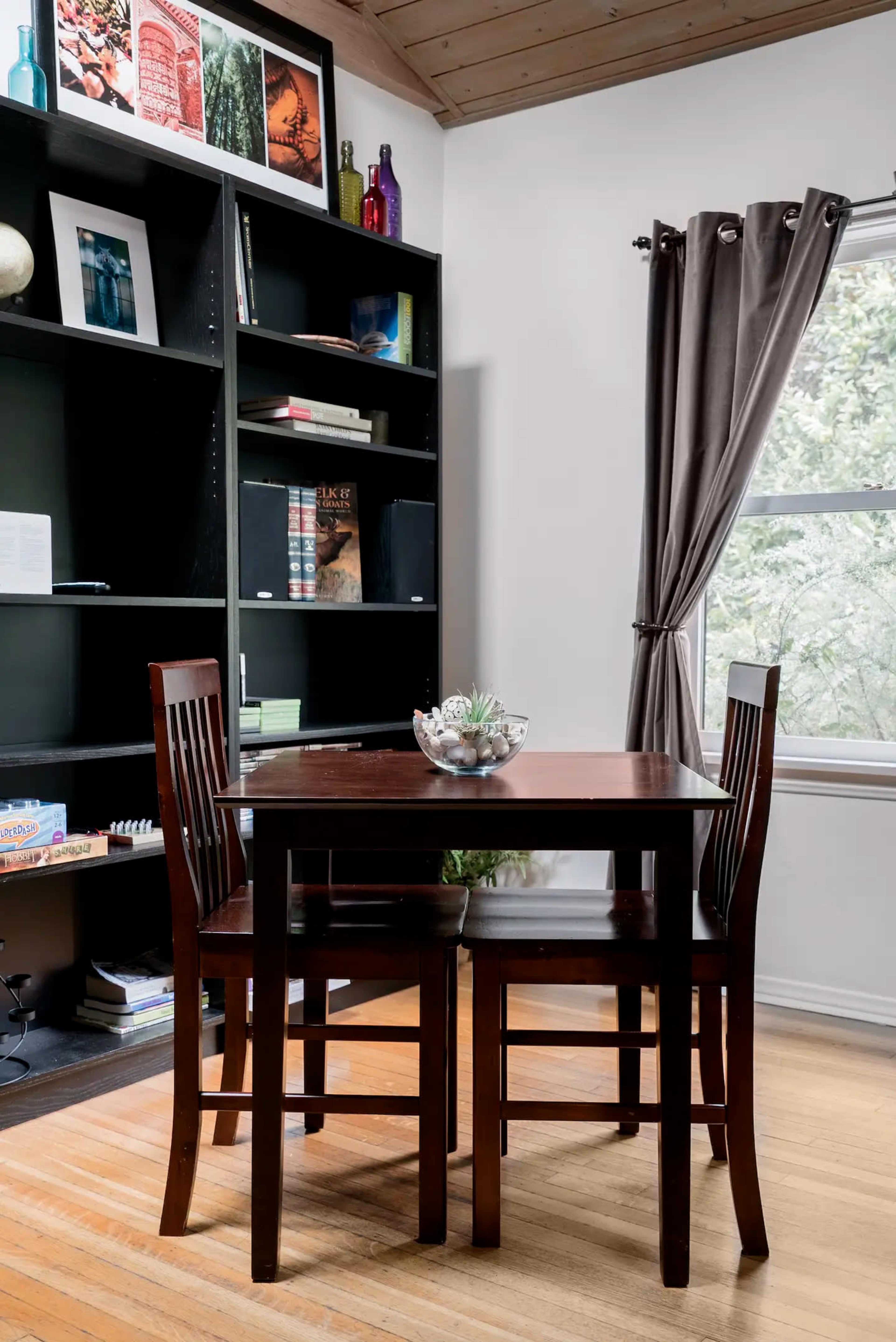 A small wooden dining table with two chairs is set against a black bookshelf filled with books and decorative items, beside a window with gray curtains.