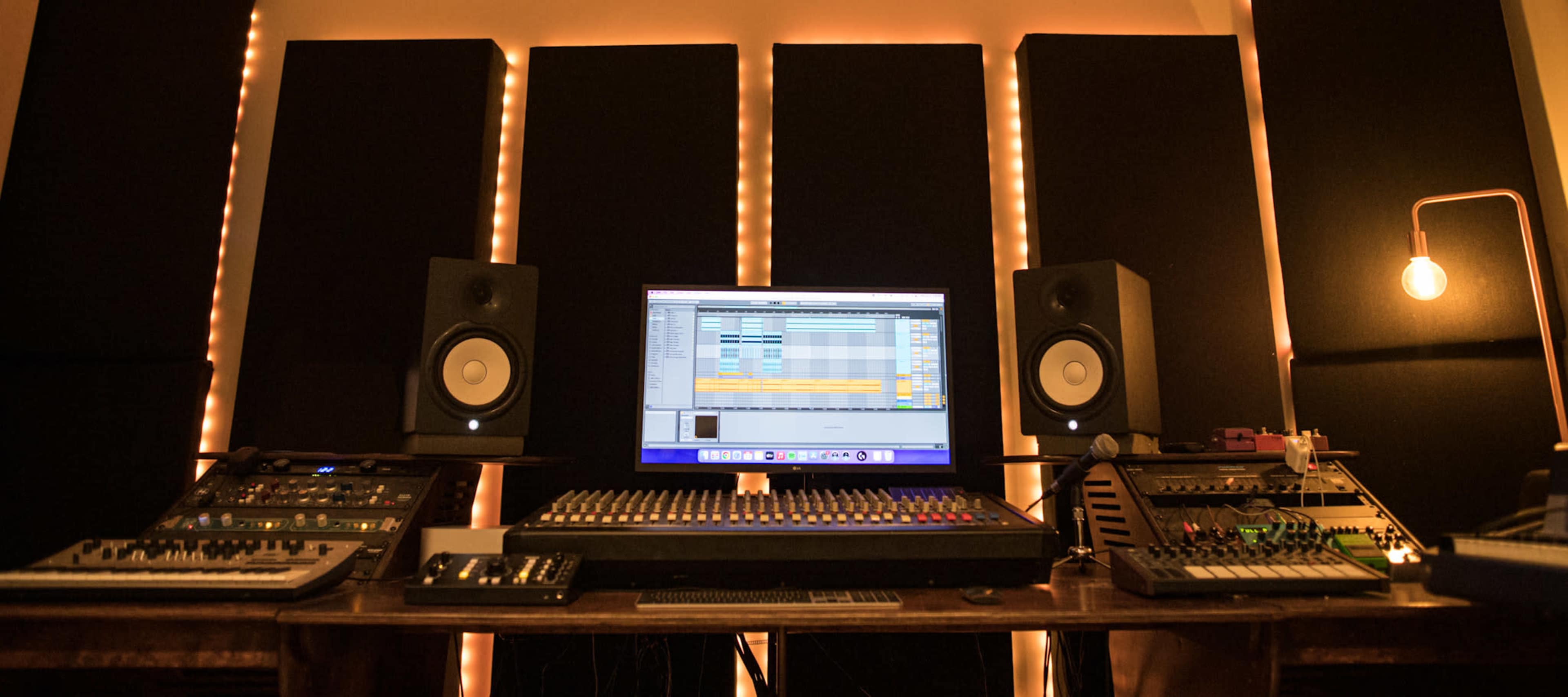 A music production studio setup with a computer monitor, audio speakers, a mixing console, and various electronic music devices arranged on a wooden table.