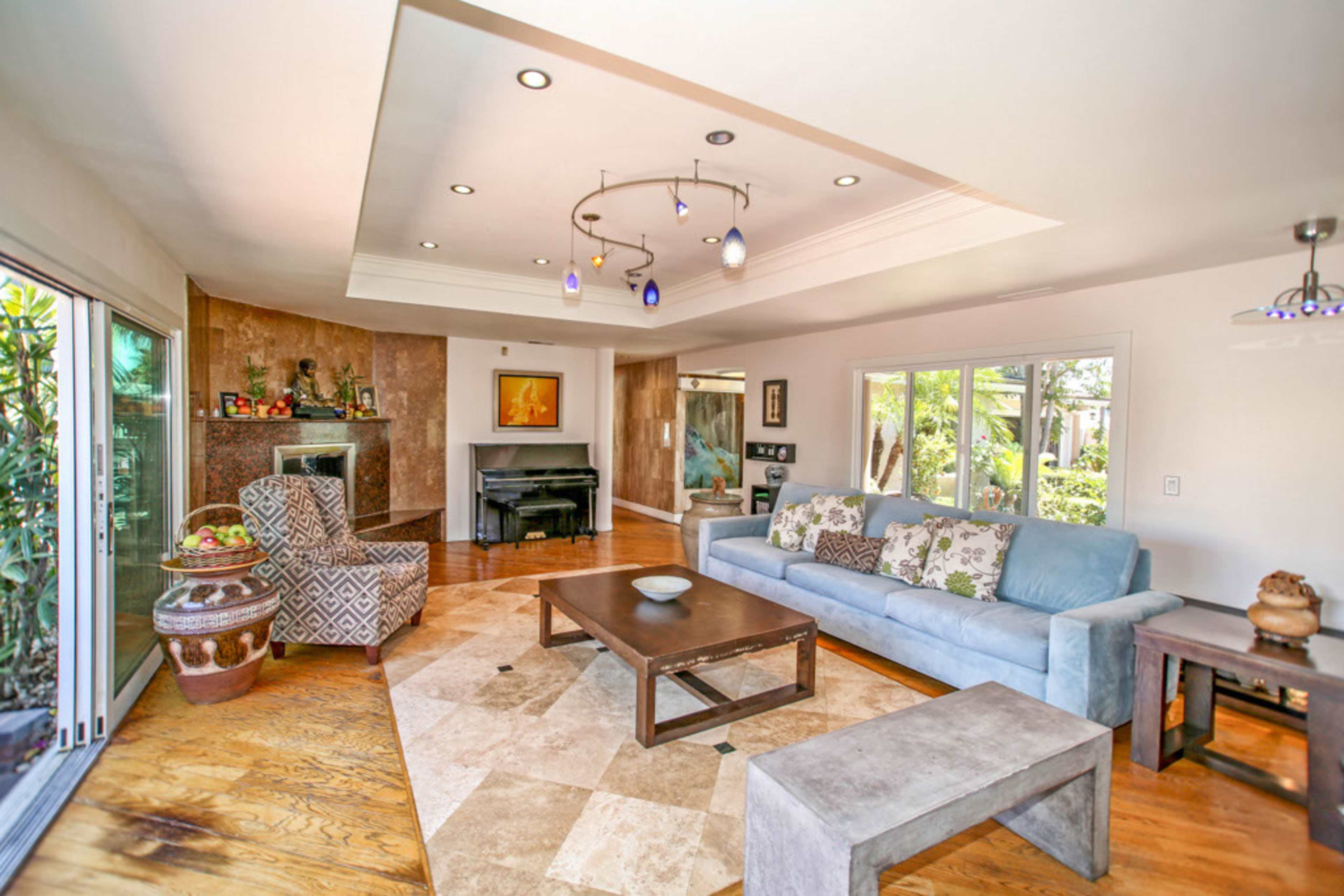 The image shows a spacious living room with a blue couch, a coffee table, and large windows overlooking a garden.