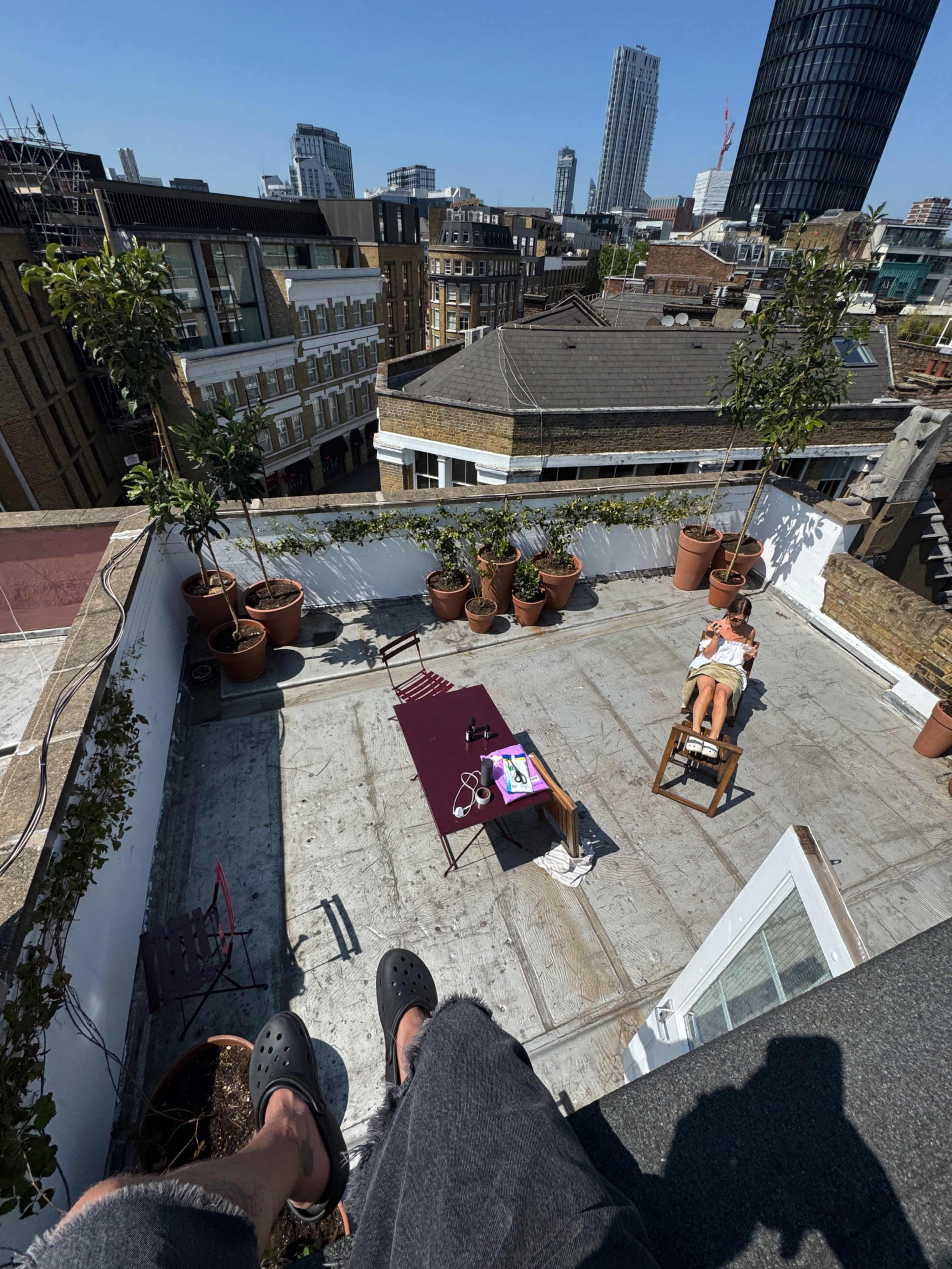The image shows a rooftop terrace in an urban setting with a person lounging on a deck chair surrounded by potted plants and a table displaying a device.