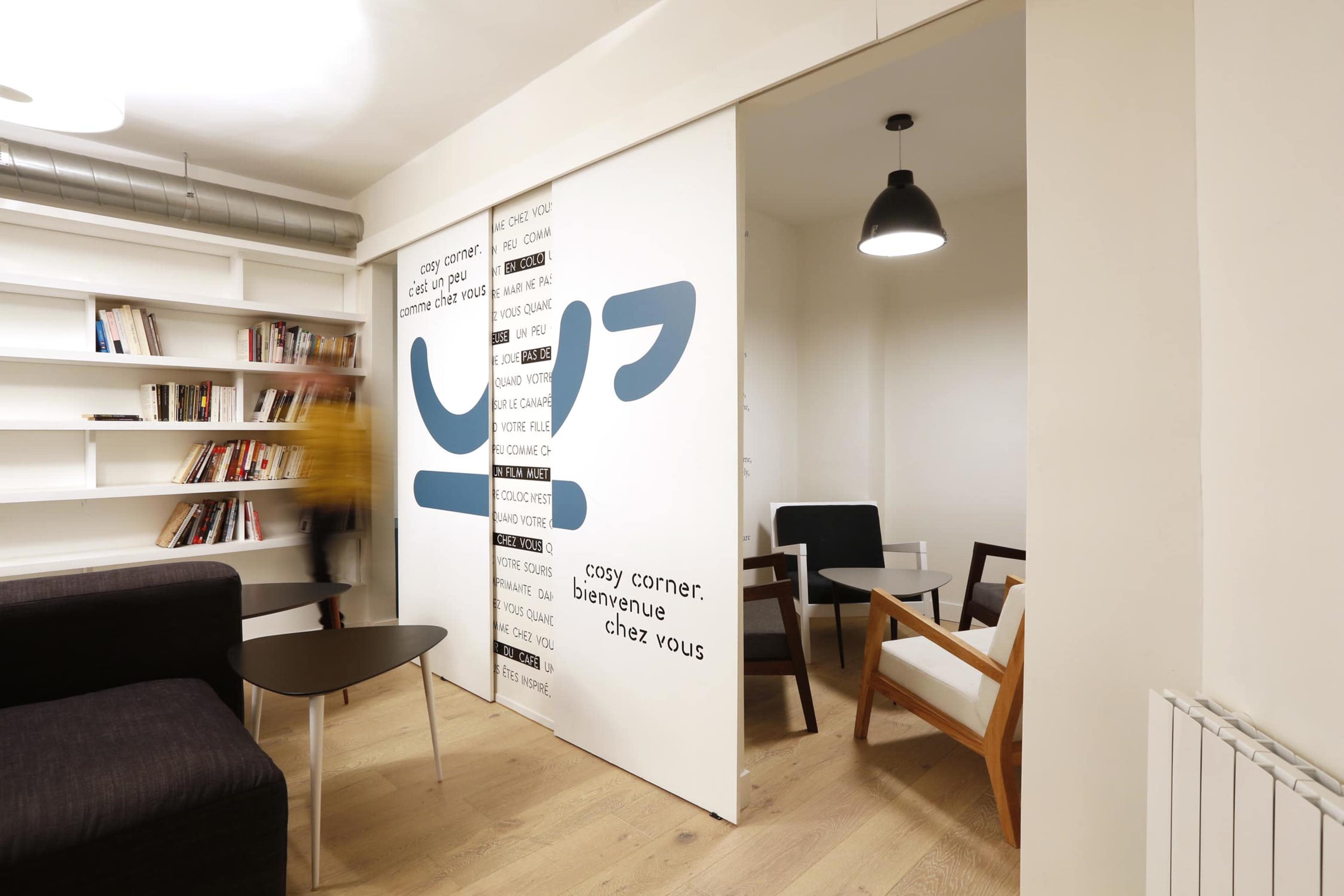 The image shows a modern, minimalist office space with a sliding partition featuring text, a small table, and a combination of chairs.