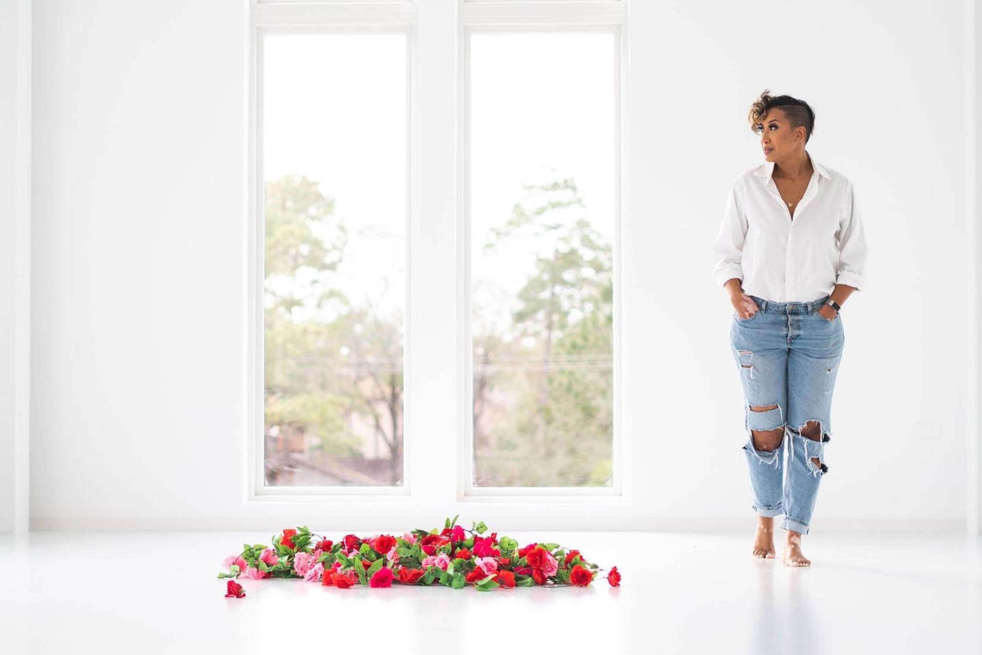 A person stands beside a heart-shaped arrangement of flowers on a bright, empty floor with large windows.