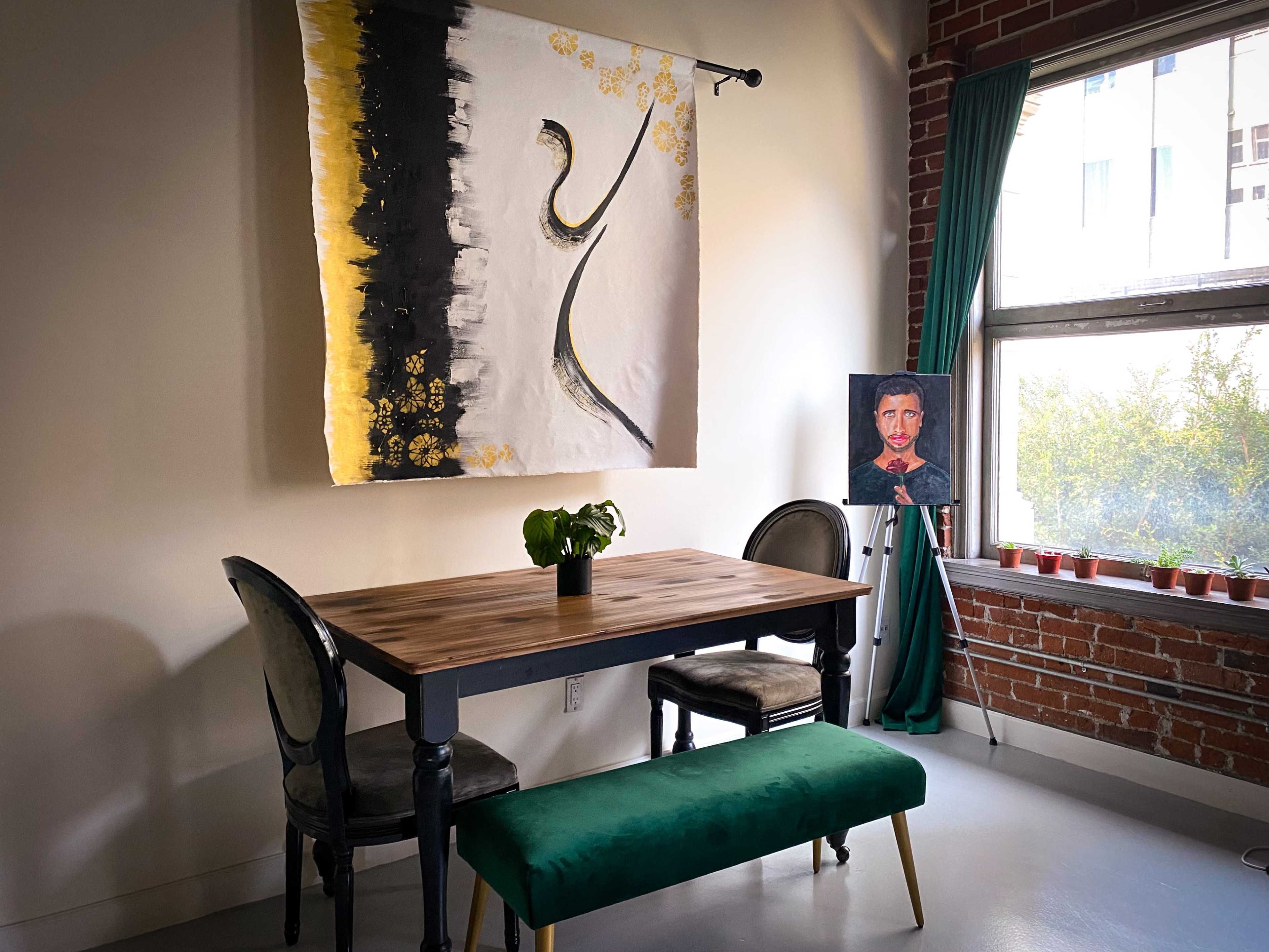 A wooden table and green bench are positioned in a room with a large wall hanging and a framed portrait next to a window.