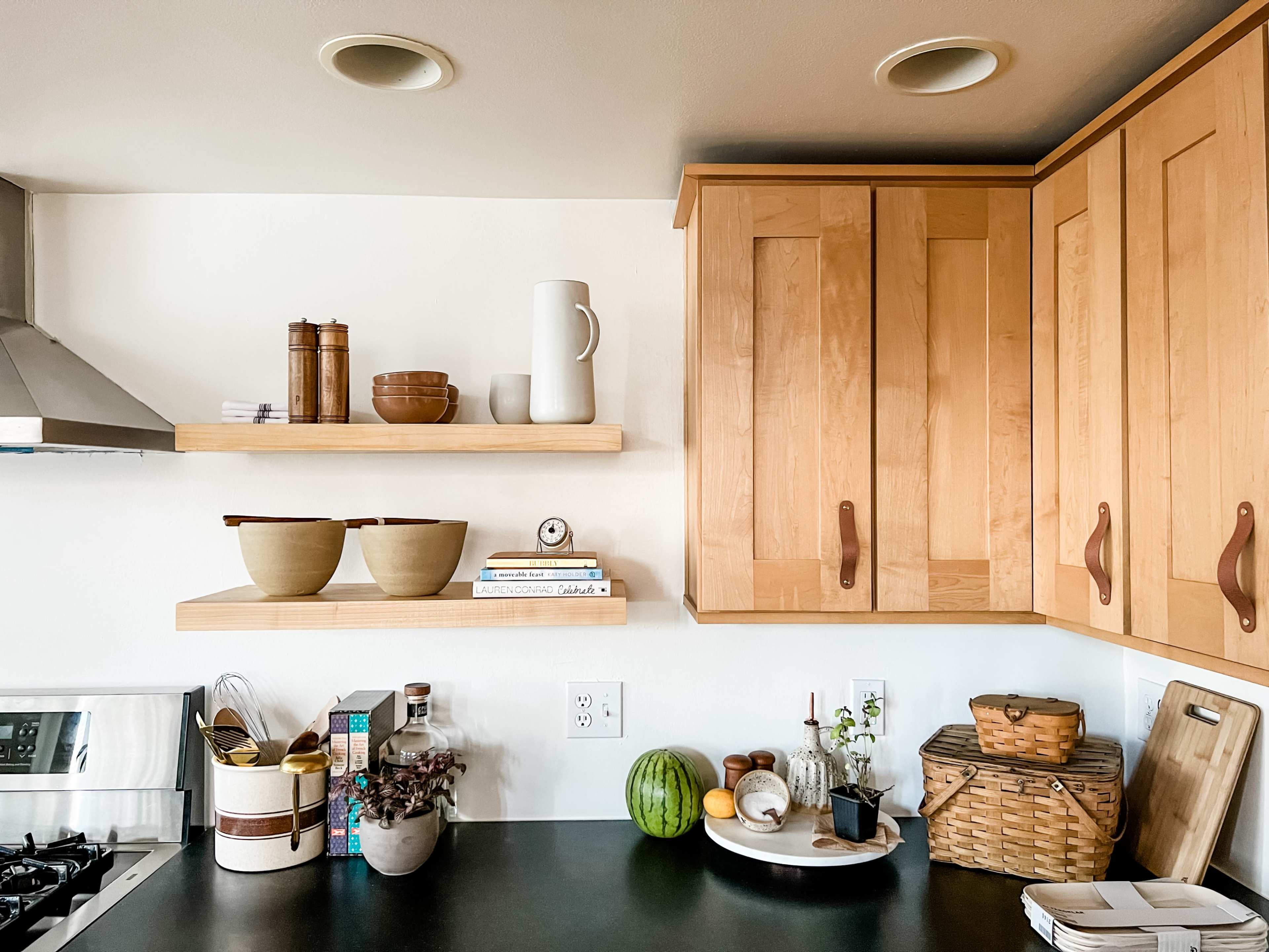 A kitchen with wooden shelves displaying bowls, a pitcher, books, and various decorative items above a black countertop with cooking utensils and a watermelon.