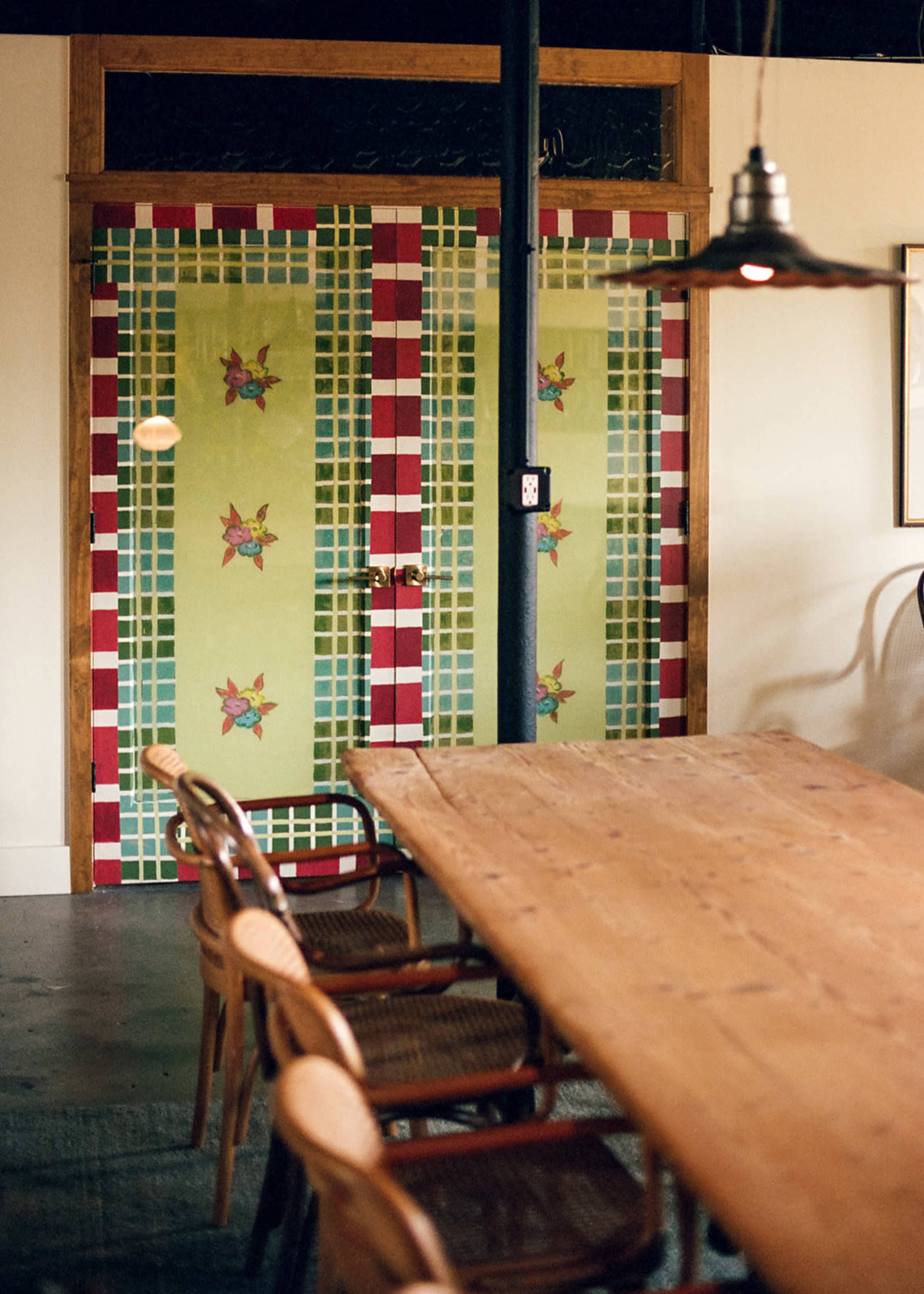 A colorful tiled door with floral designs is set against a light-colored wall within a dining area featuring a large wooden table and chairs.