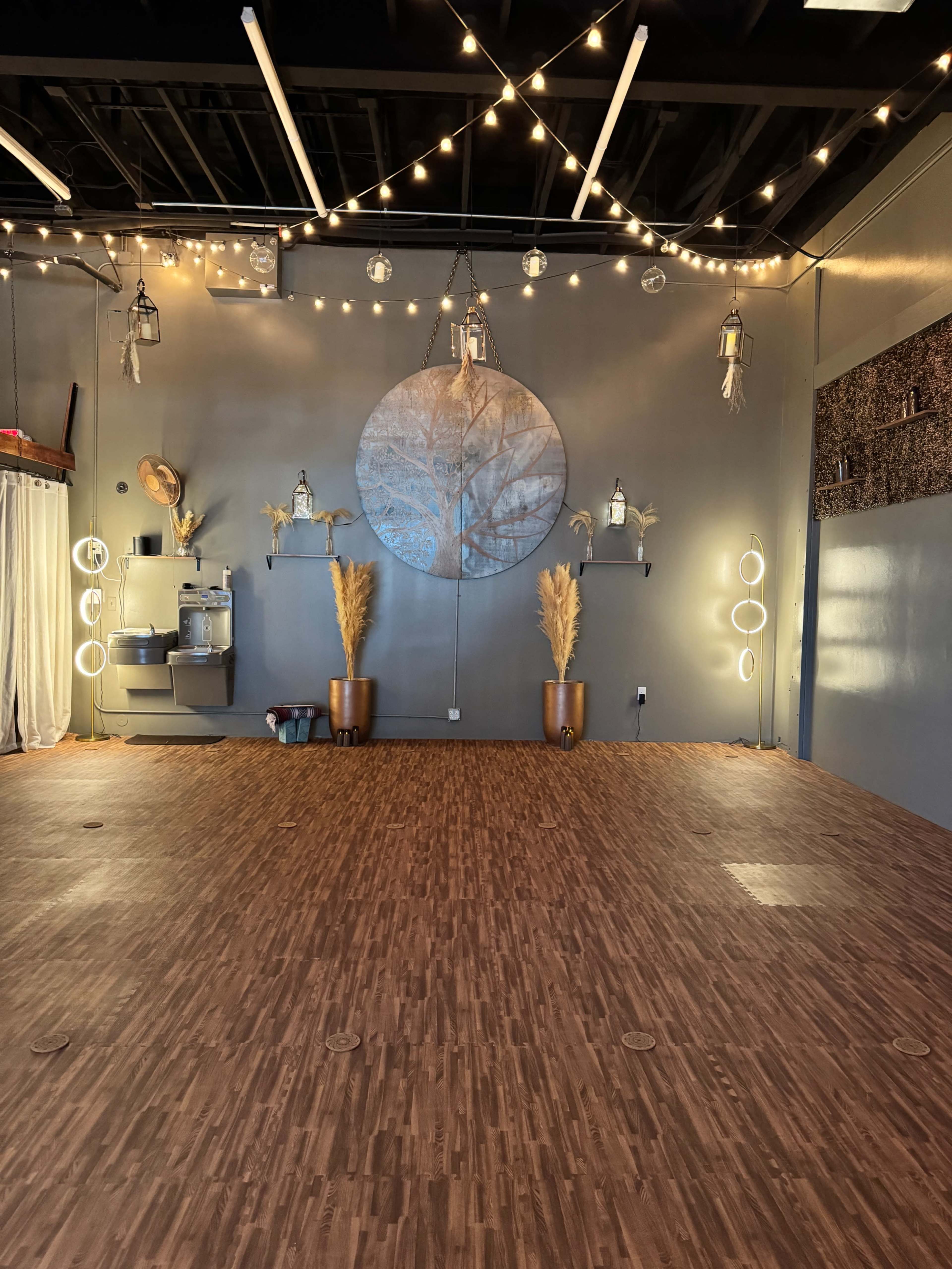 The image shows a spacious interior with wooden flooring, decorative wall hangings, and various lighting fixtures, including string lights overhead.