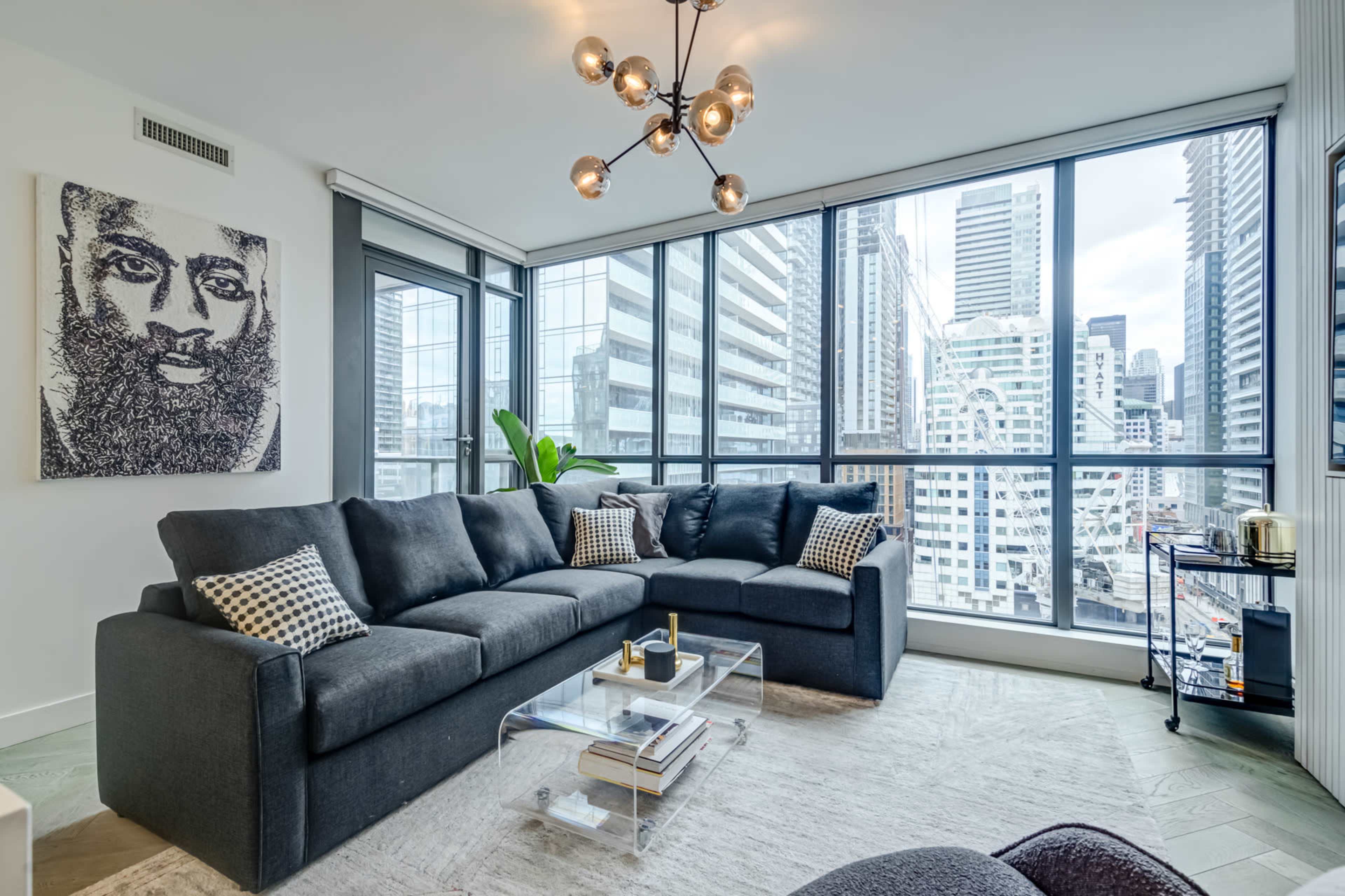 A modern living room features a large gray sectional sofa, a glass coffee table, and a view of city skyscrapers through large windows.