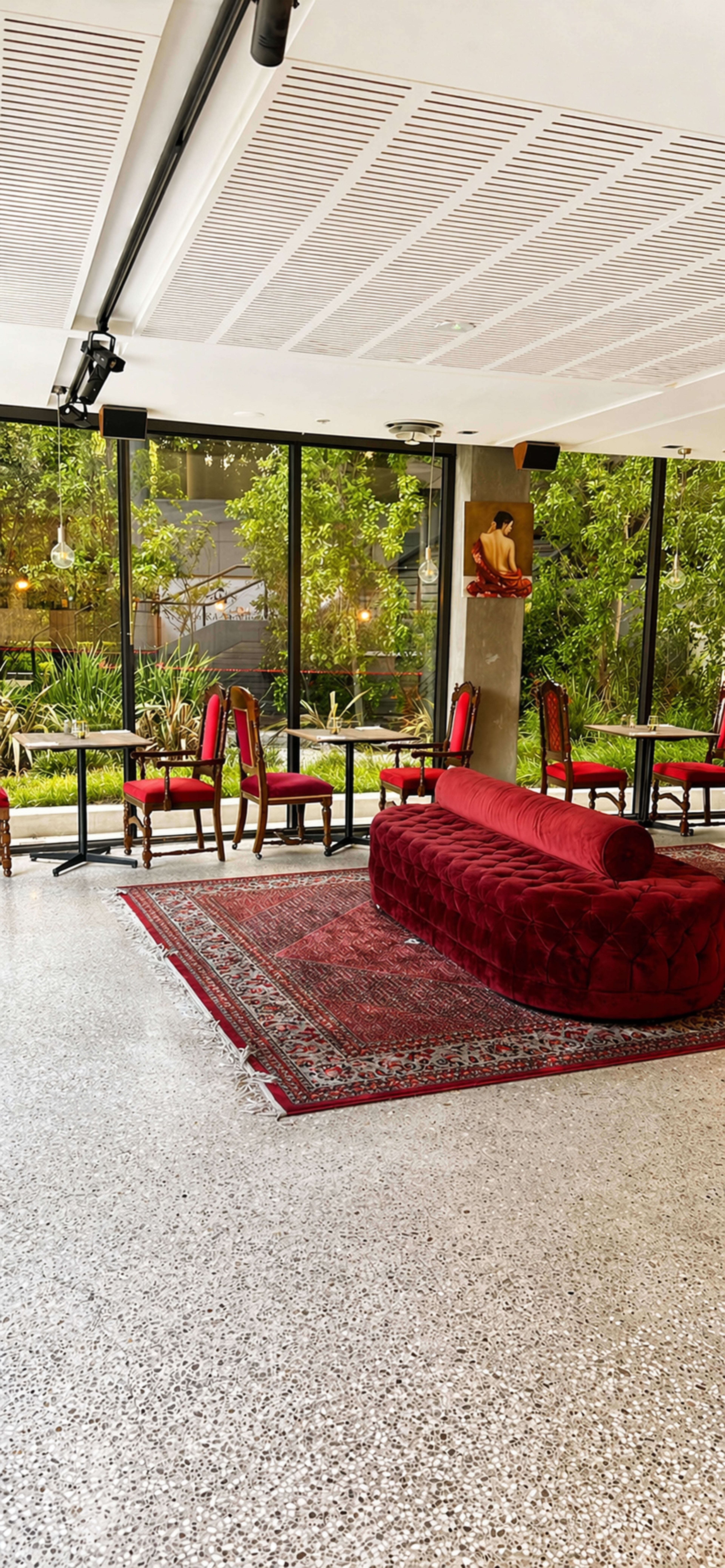 The image shows a modern interior space featuring a red upholstered sofa, a patterned rug on the polished floor, and tables surrounded by chairs near large glass windows that overlook greenery.