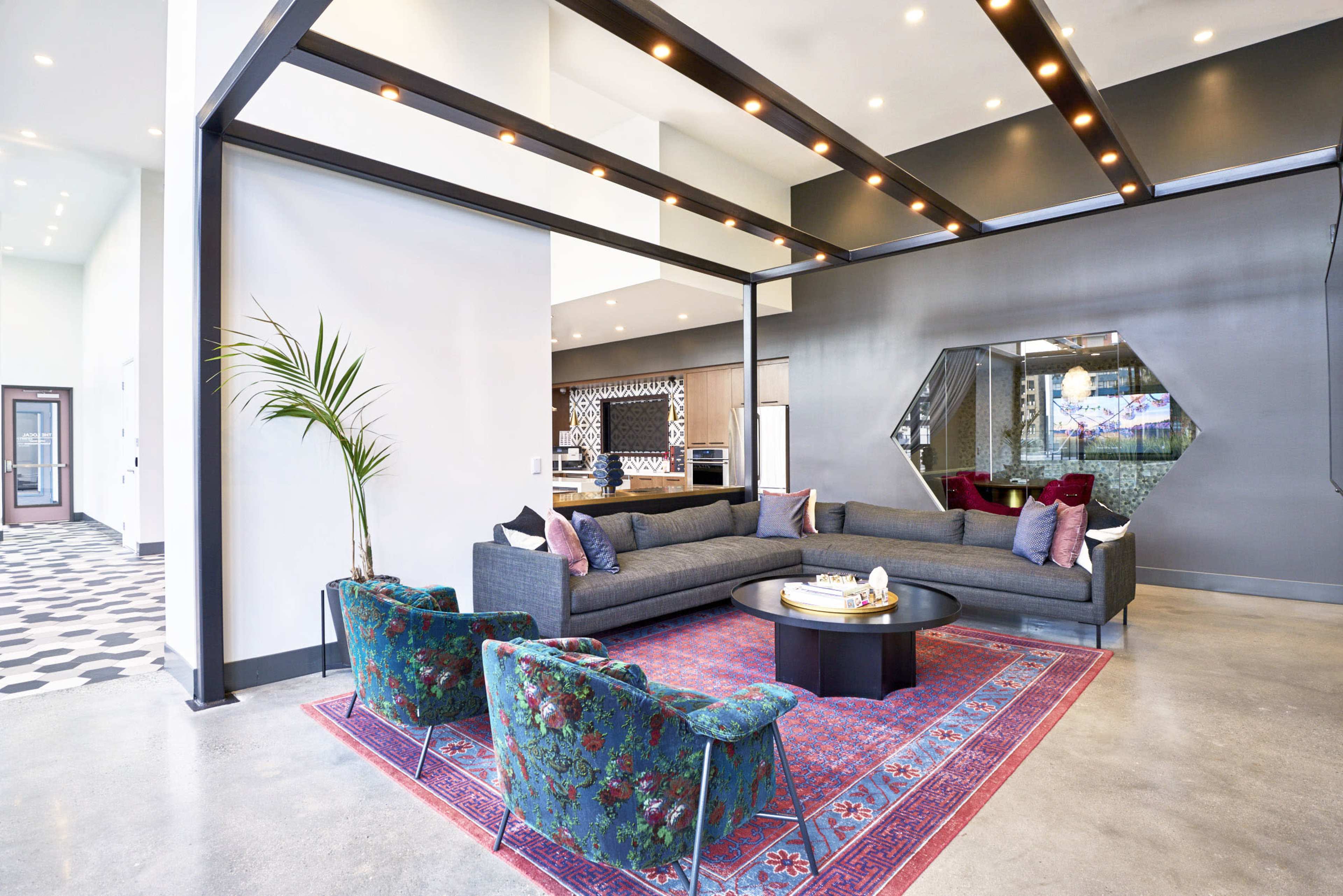 A modern living area features a sectional sofa, two patterned armchairs, and a round coffee table on a decorative rug, with a kitchen visible in the background.