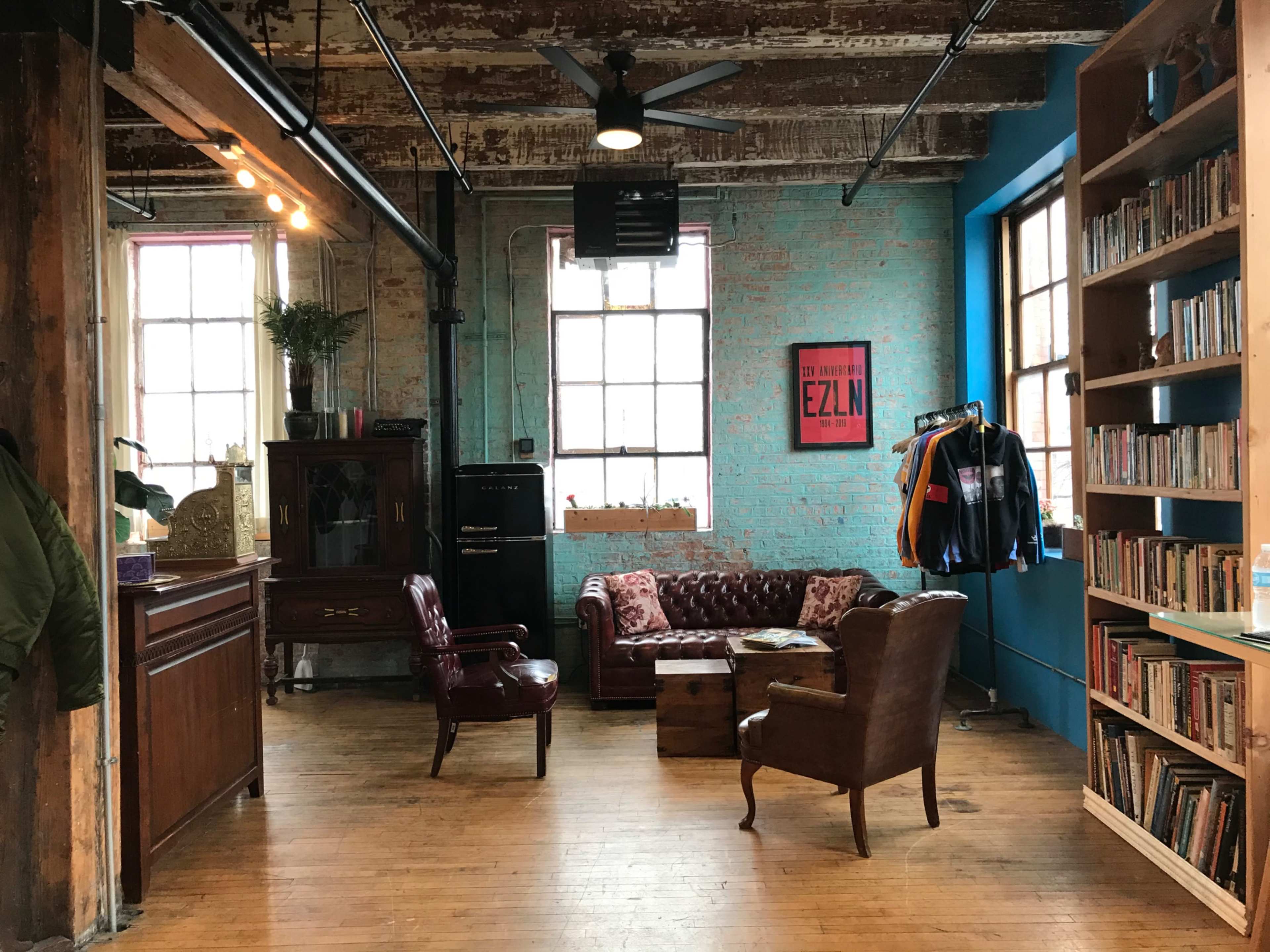 A spacious room with exposed brick walls, wooden floors, and a seating area featuring two armchairs and a coffee table, along with a bookshelf and a vintage cabinet.