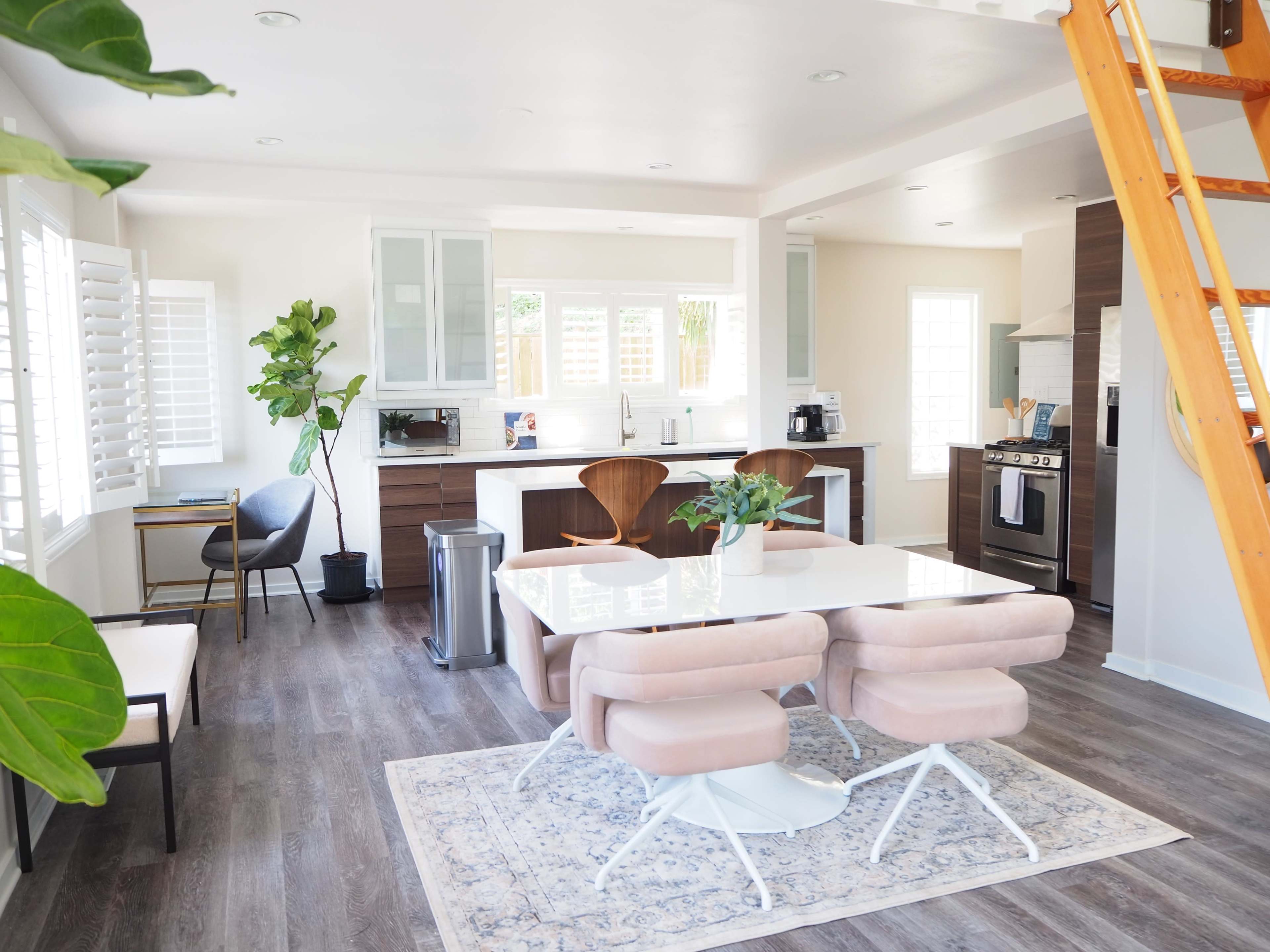 A modern kitchen and dining area with a white table, pink chairs, and a wooden ladder leading to a loft space.
