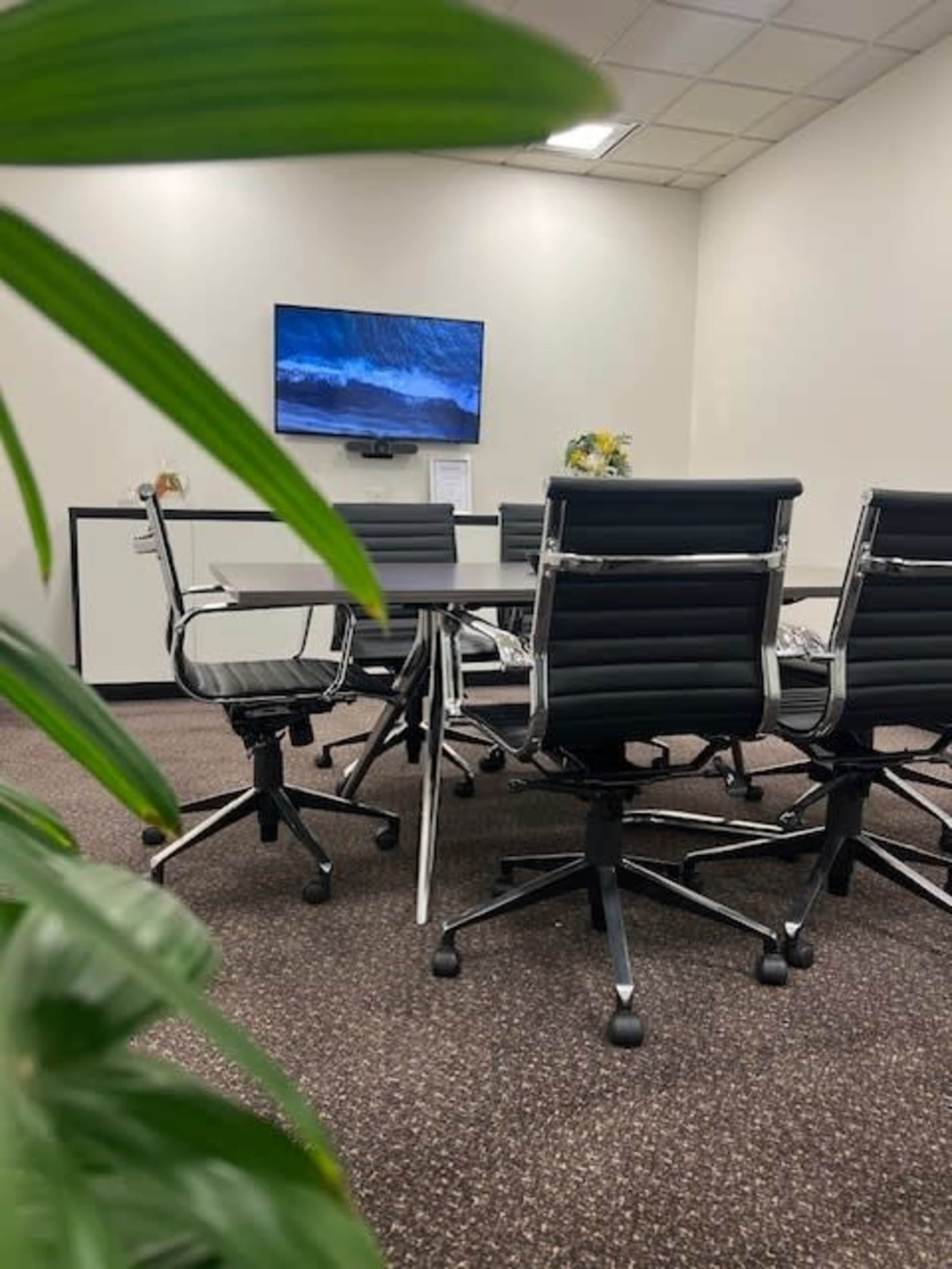A modern conference room features a large table surrounded by ergonomic chairs, with a television mounted on the wall displaying a scenic image.