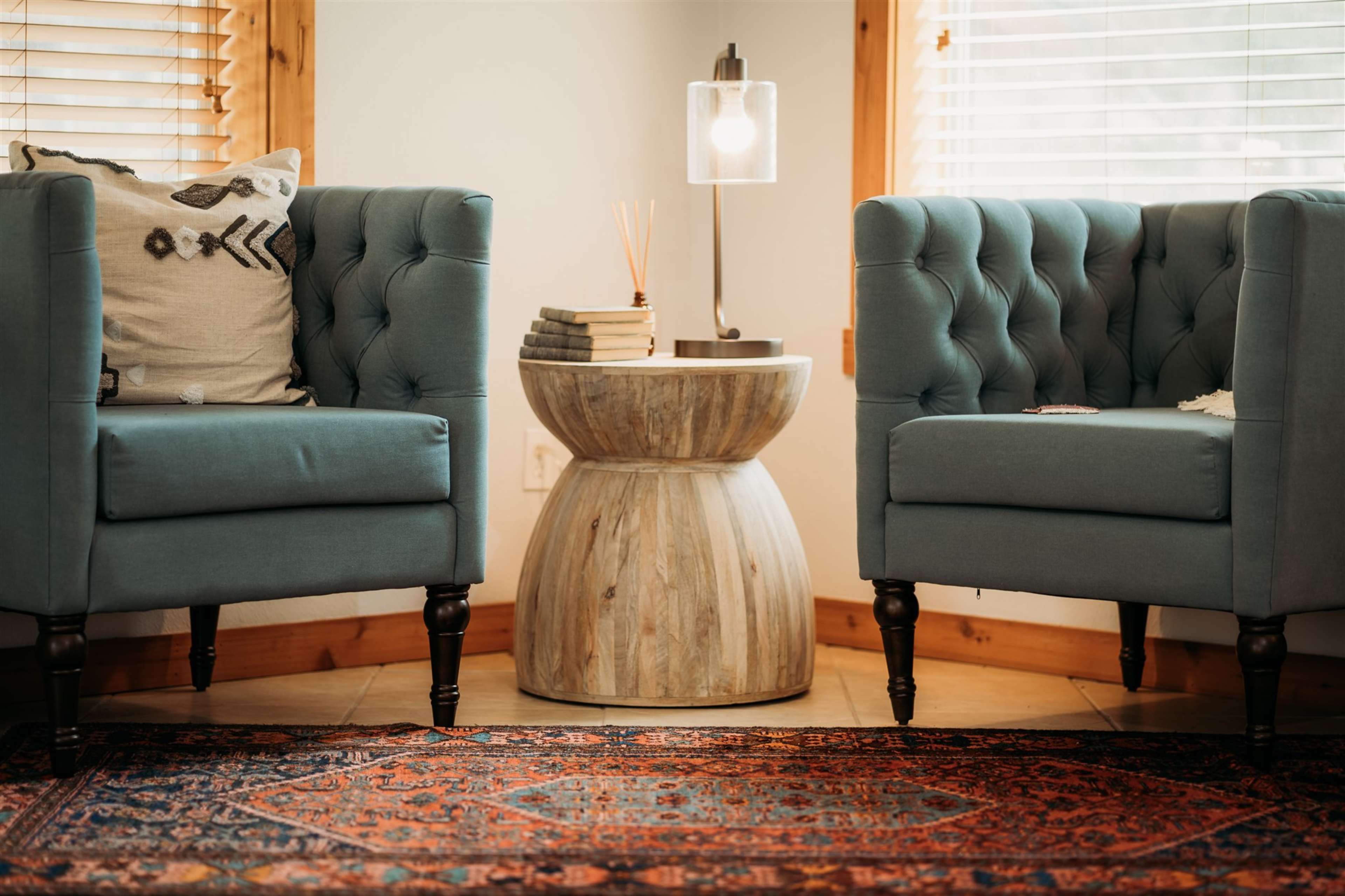 Two tufted blue armchairs flanking a round wooden side table, with a lamp and decorative books on top, set on a patterned rug in a well-lit room.