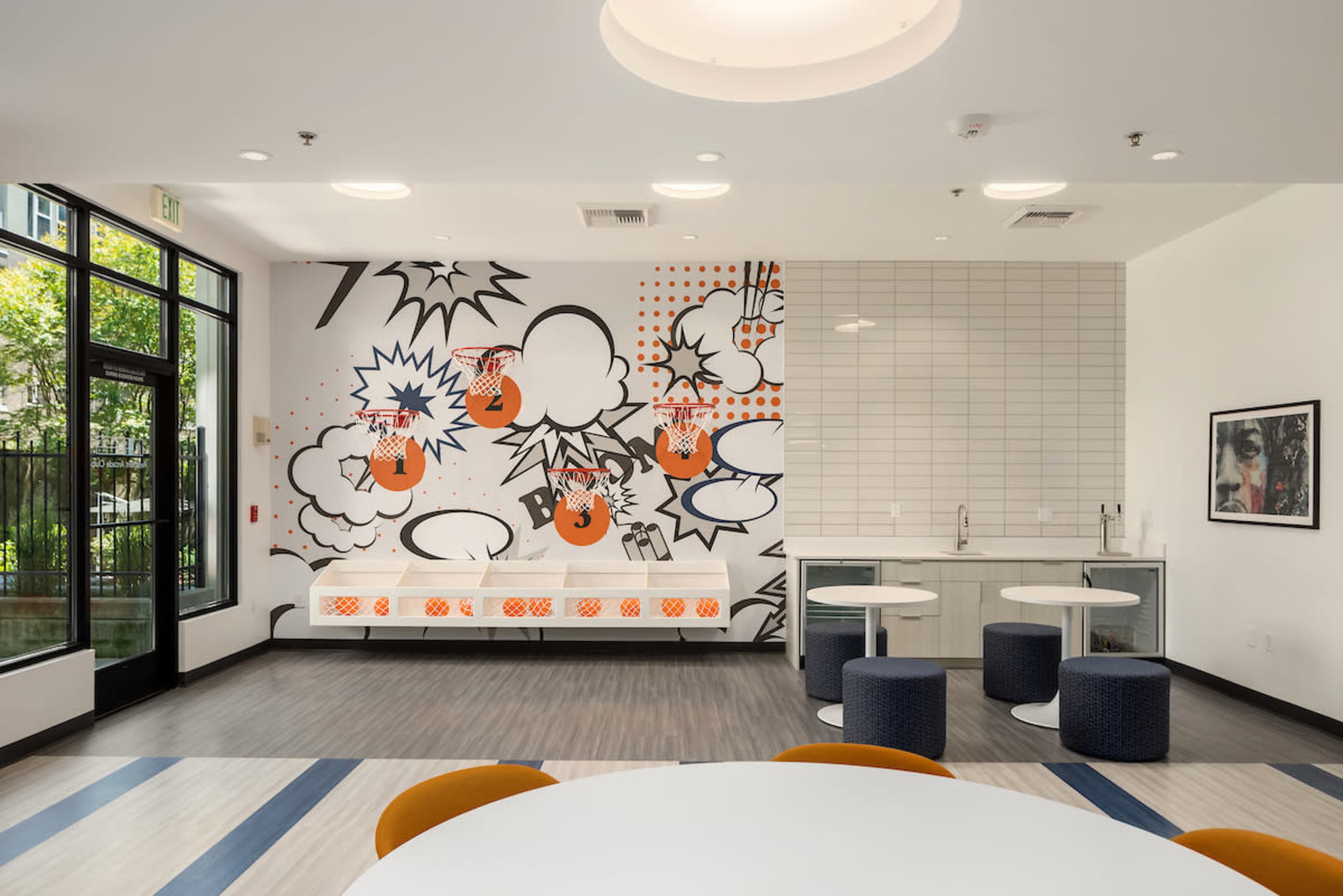 A modern communal space with a large graphic mural on one wall, a kitchenette area, and various seating options, including round tables and stools.