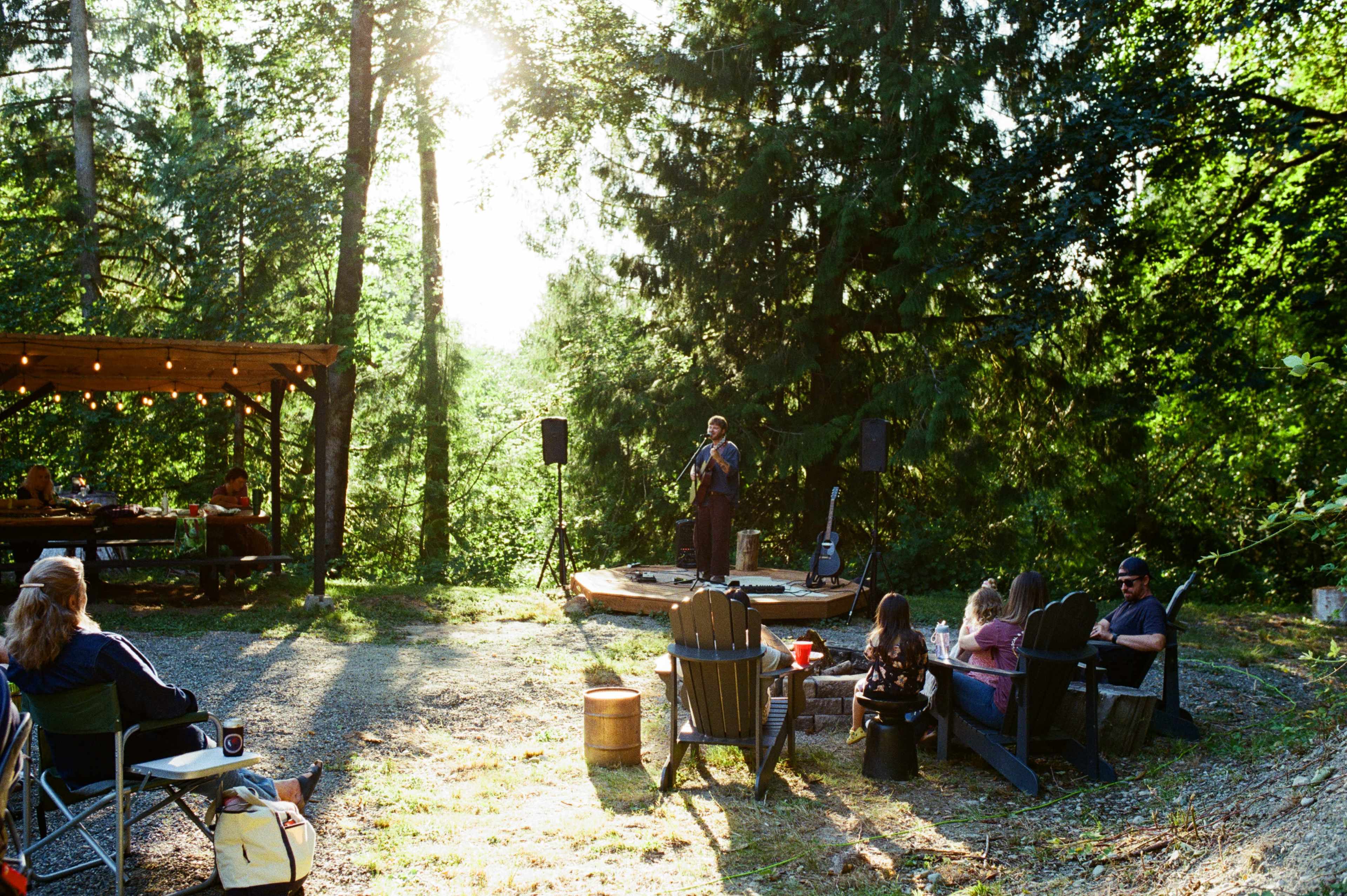 A musician performs on a circular stage surrounded by an audience seated in outdoor chairs amidst a forested setting.
