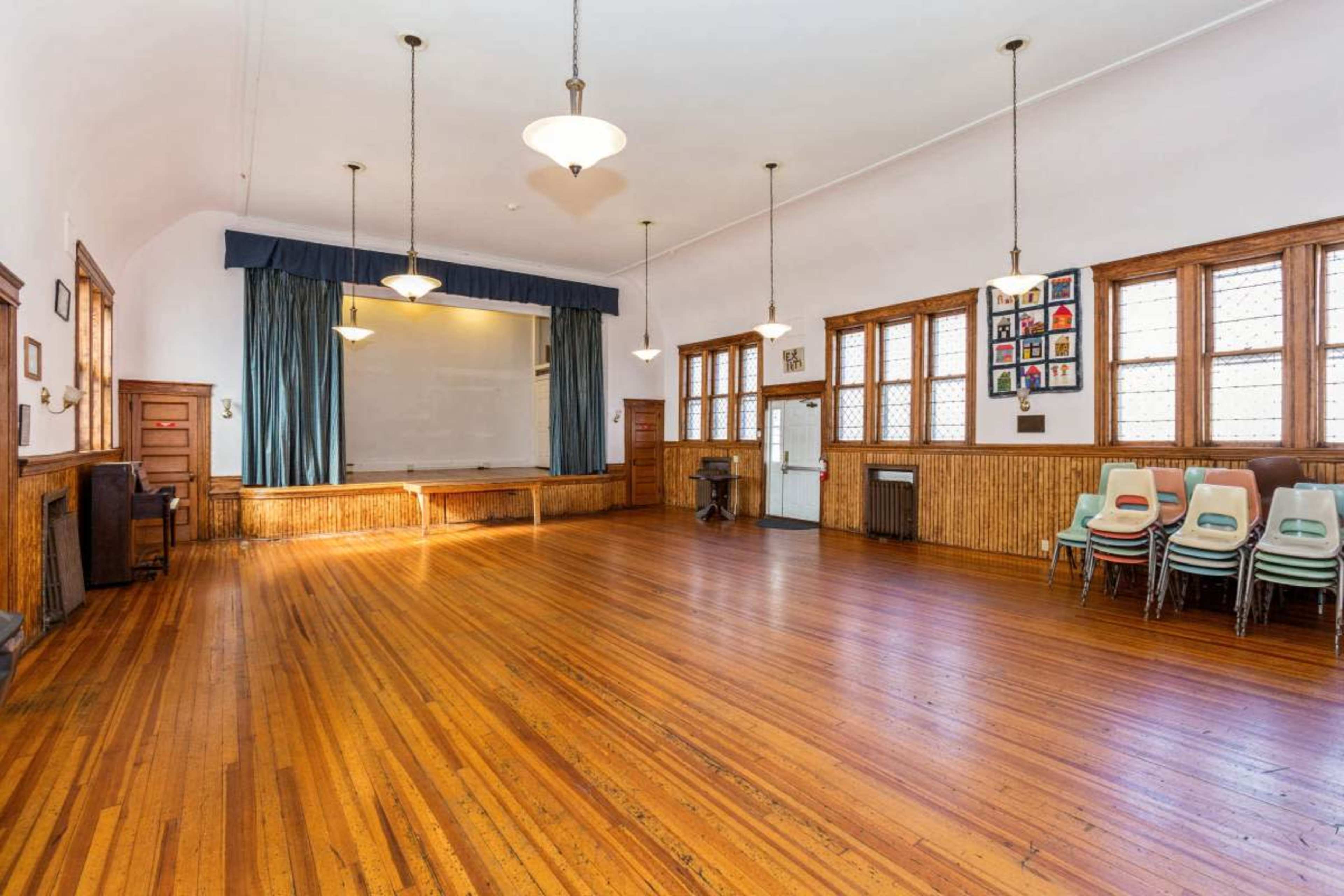 The image shows a spacious, wooden-floored hall with rows of stacked chairs against a wall and a stage at the front.