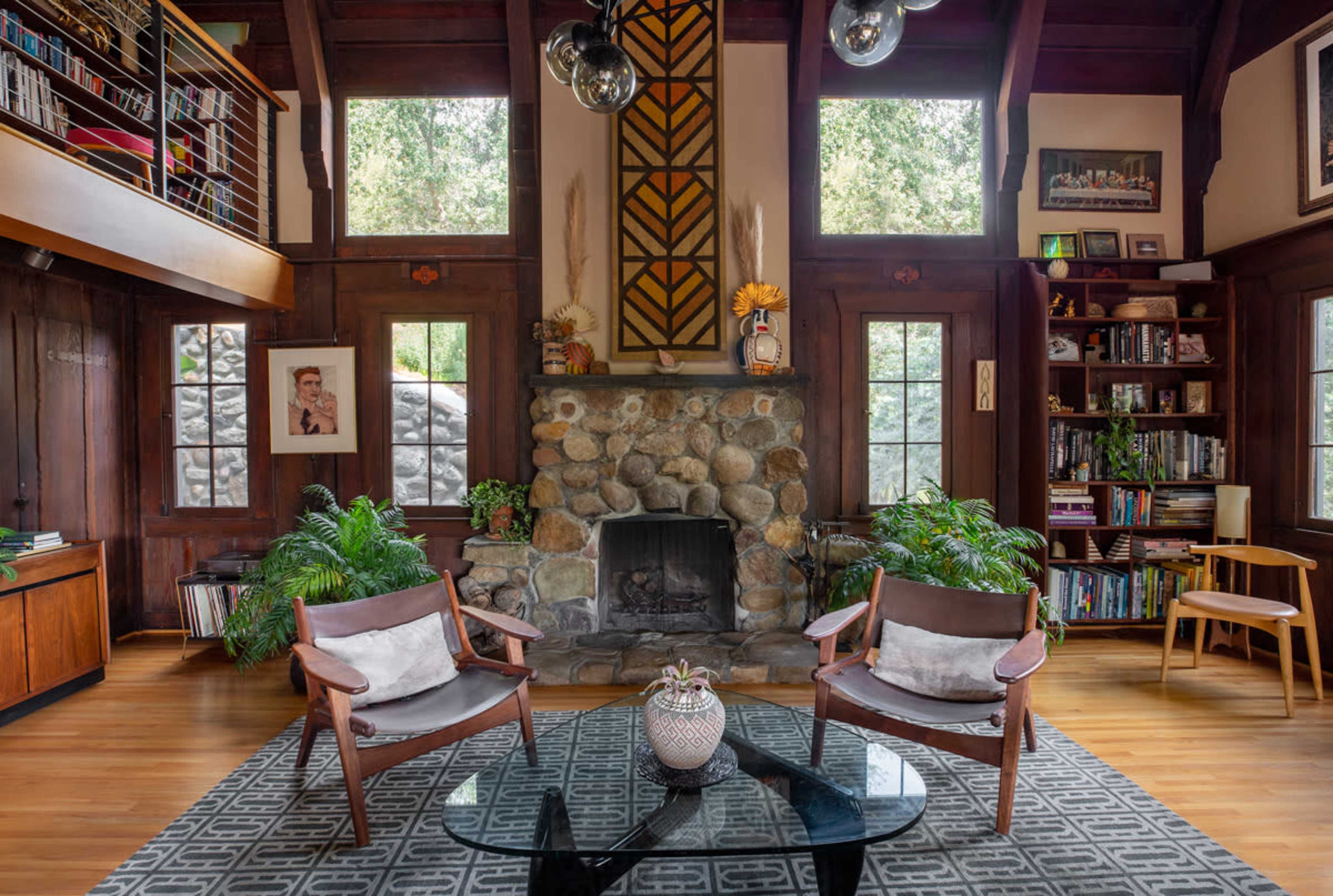 A cozy living room featuring a stone fireplace, two wooden chairs, and a bookshelf filled with books, surrounded by large windows and plants.