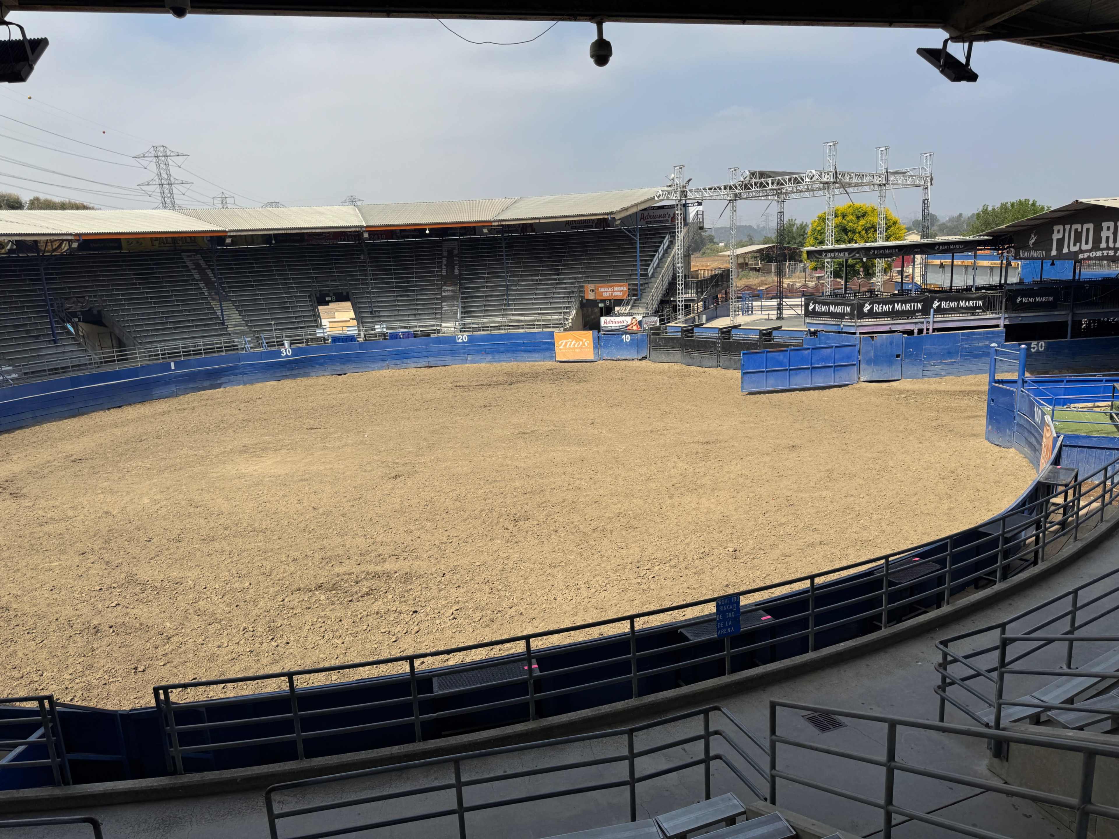 The image shows an empty rodeo arena with dirt flooring and seating arranged around the perimeter.
