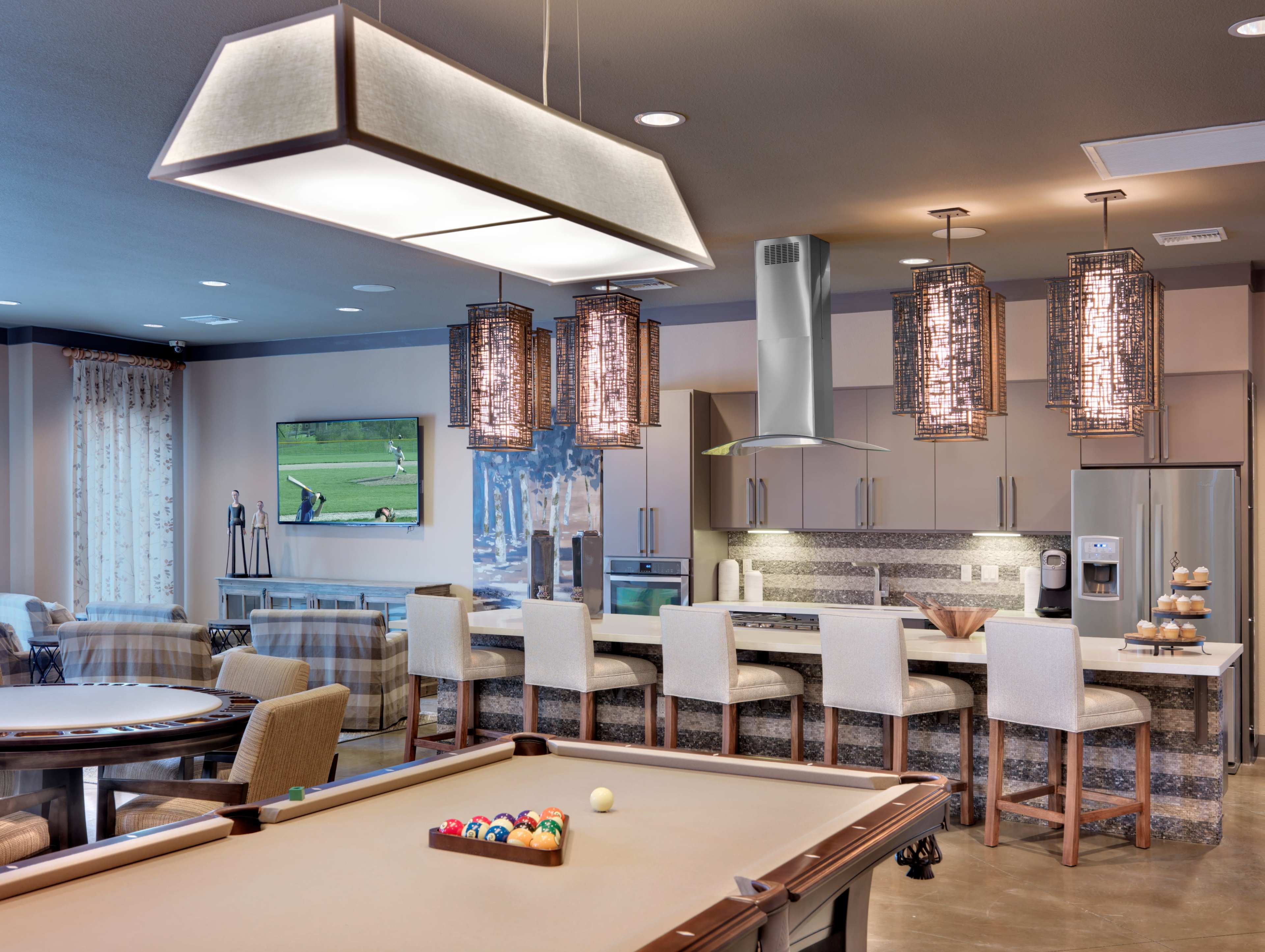 A spacious indoor recreational area features a pool table in the foreground and a kitchen bar with hanging lights in the background.
