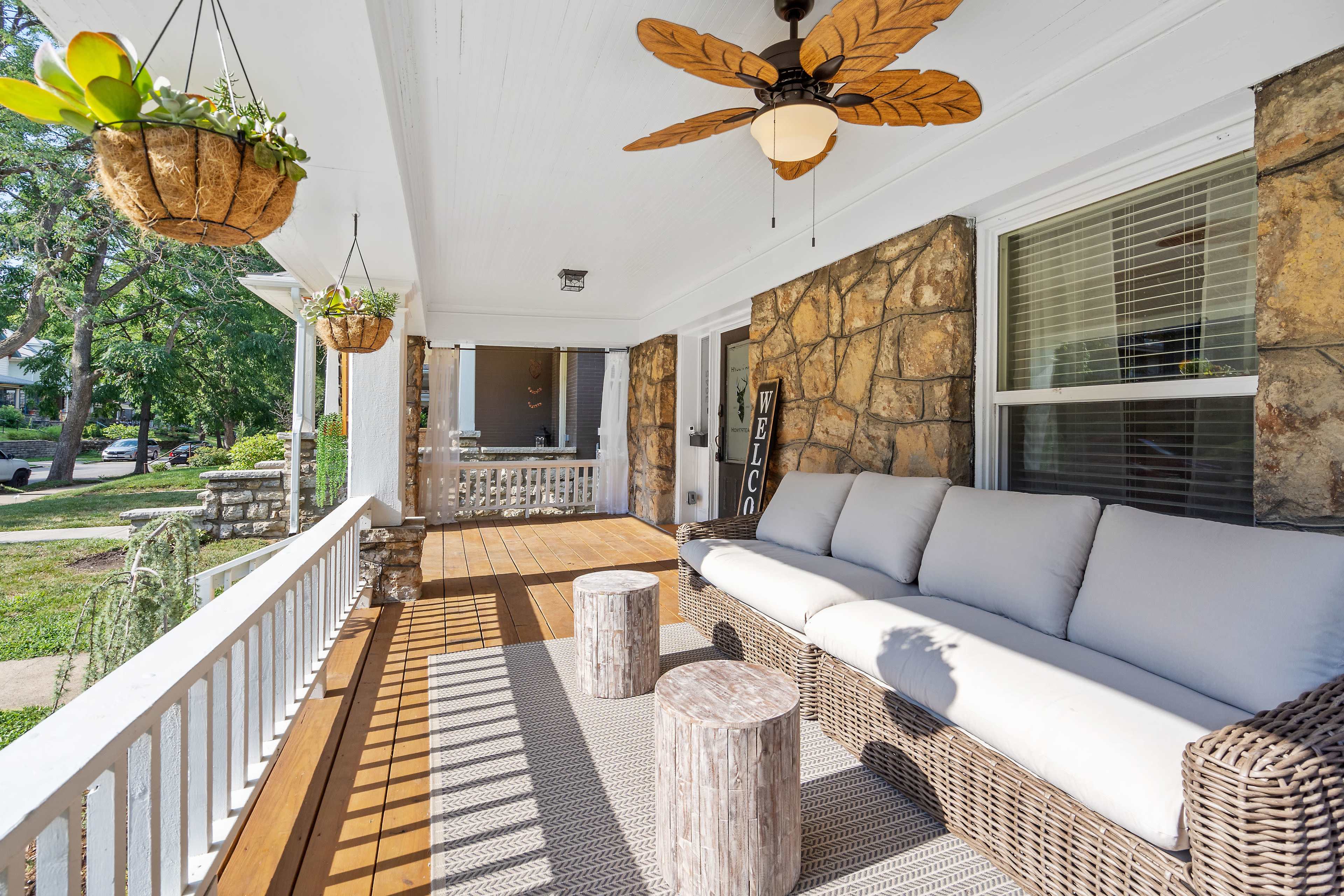 A covered porch features a wicker sofa, wooden stools, and hanging plants, with sunlight casting shadows on the floor.