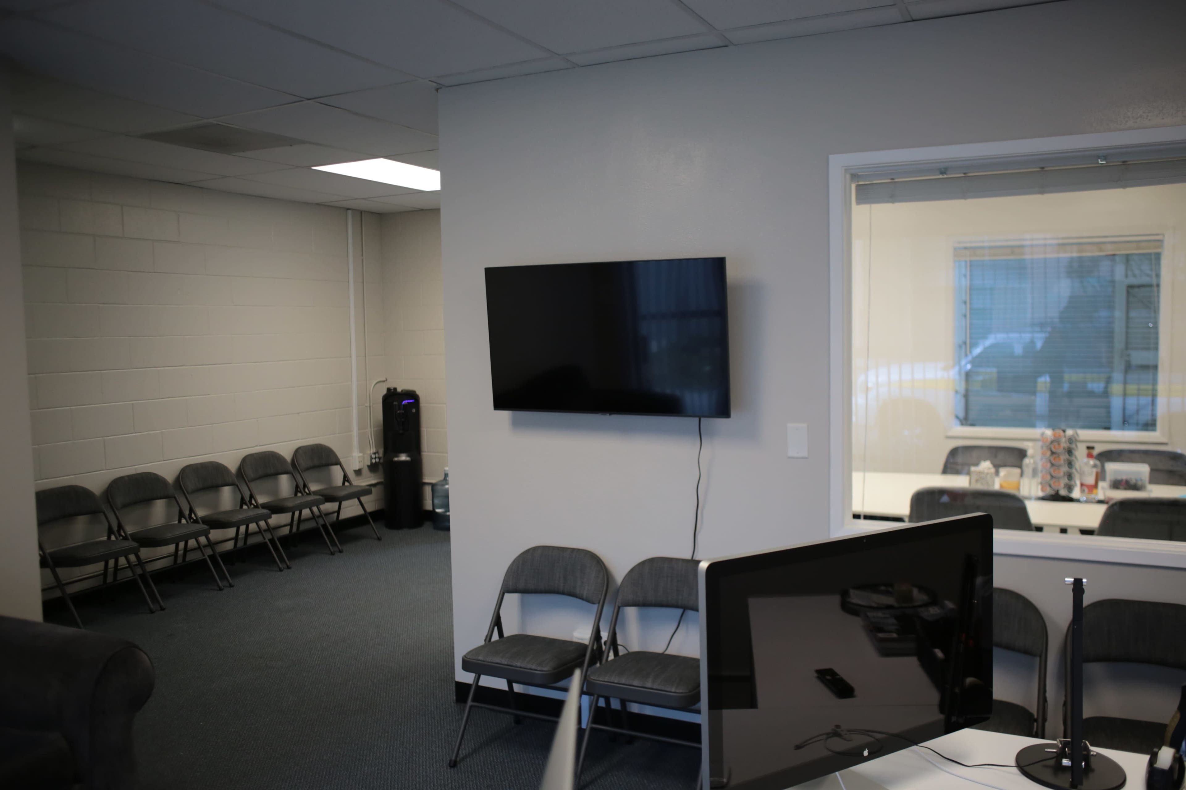 A waiting area with several gray chairs and a wall-mounted television is visible, along with an adjacent room containing a large window.