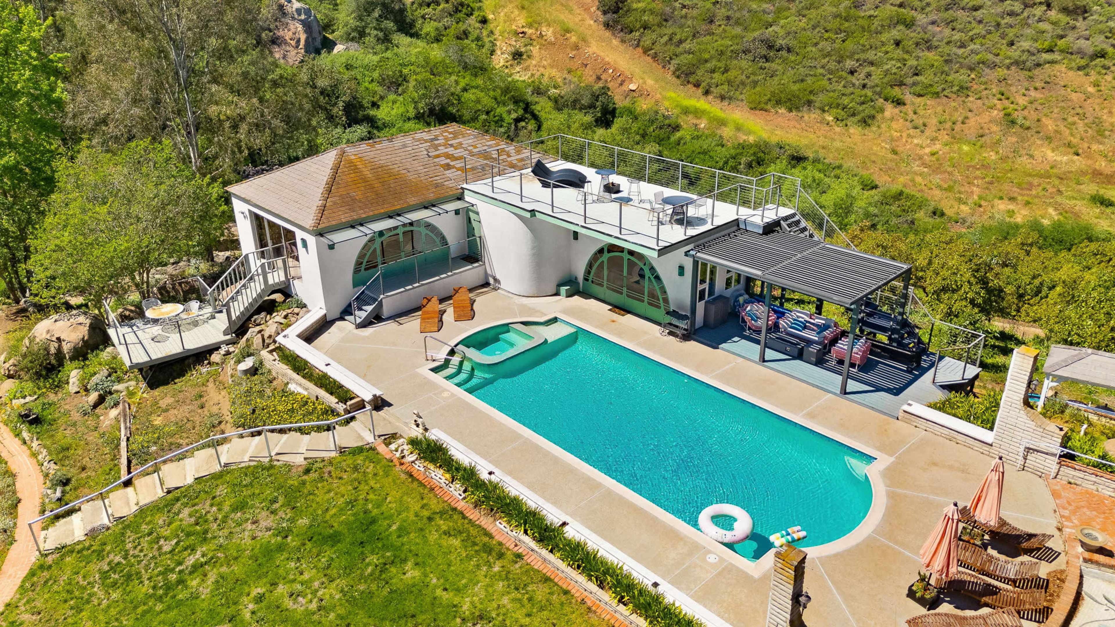 The image shows a modern house with a swimming pool, situated on a hillside surrounded by greenery.