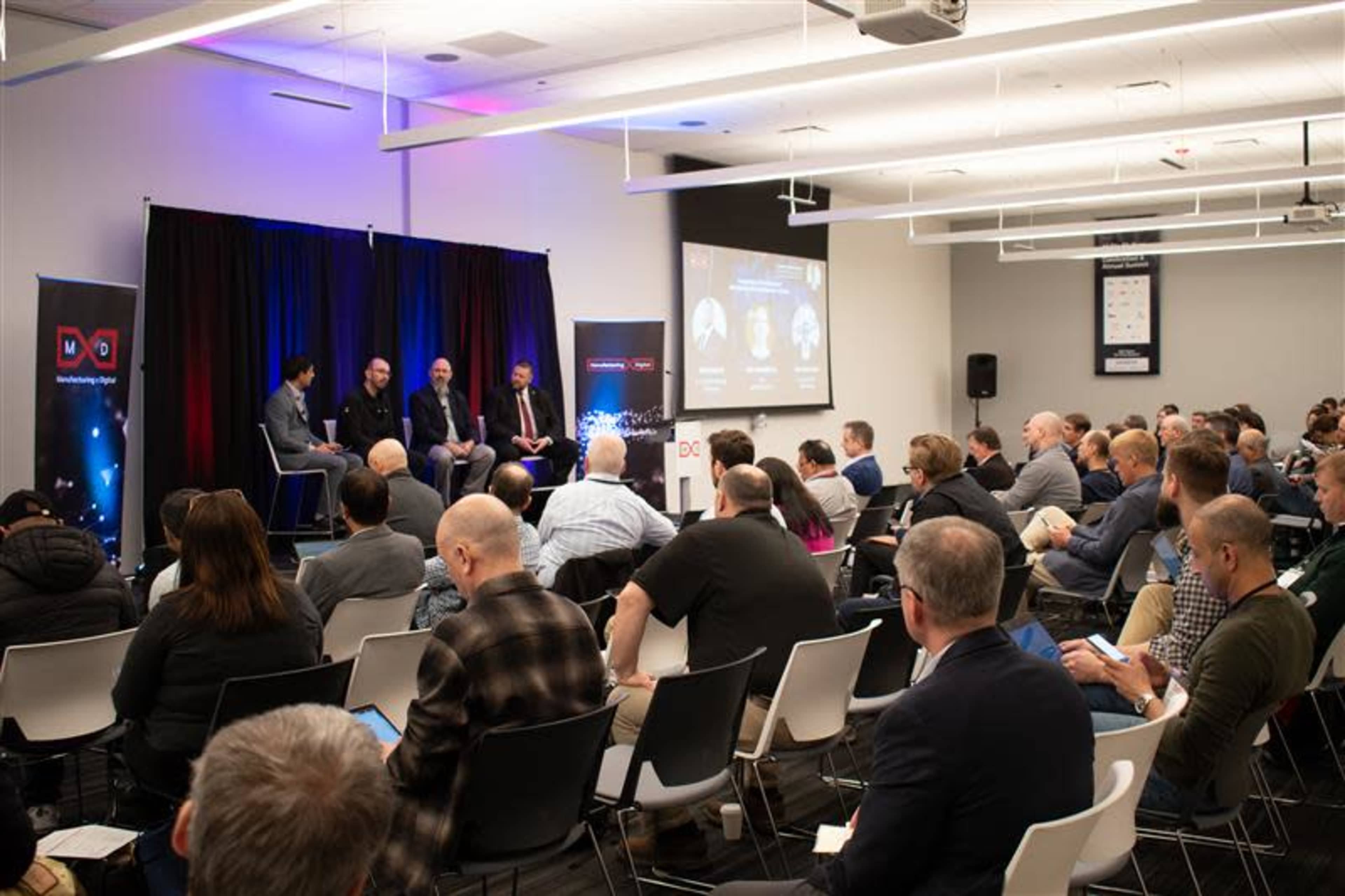 A panel discussion is taking place in a conference room filled with an audience, featuring four speakers on stage and a projector displaying information behind them.