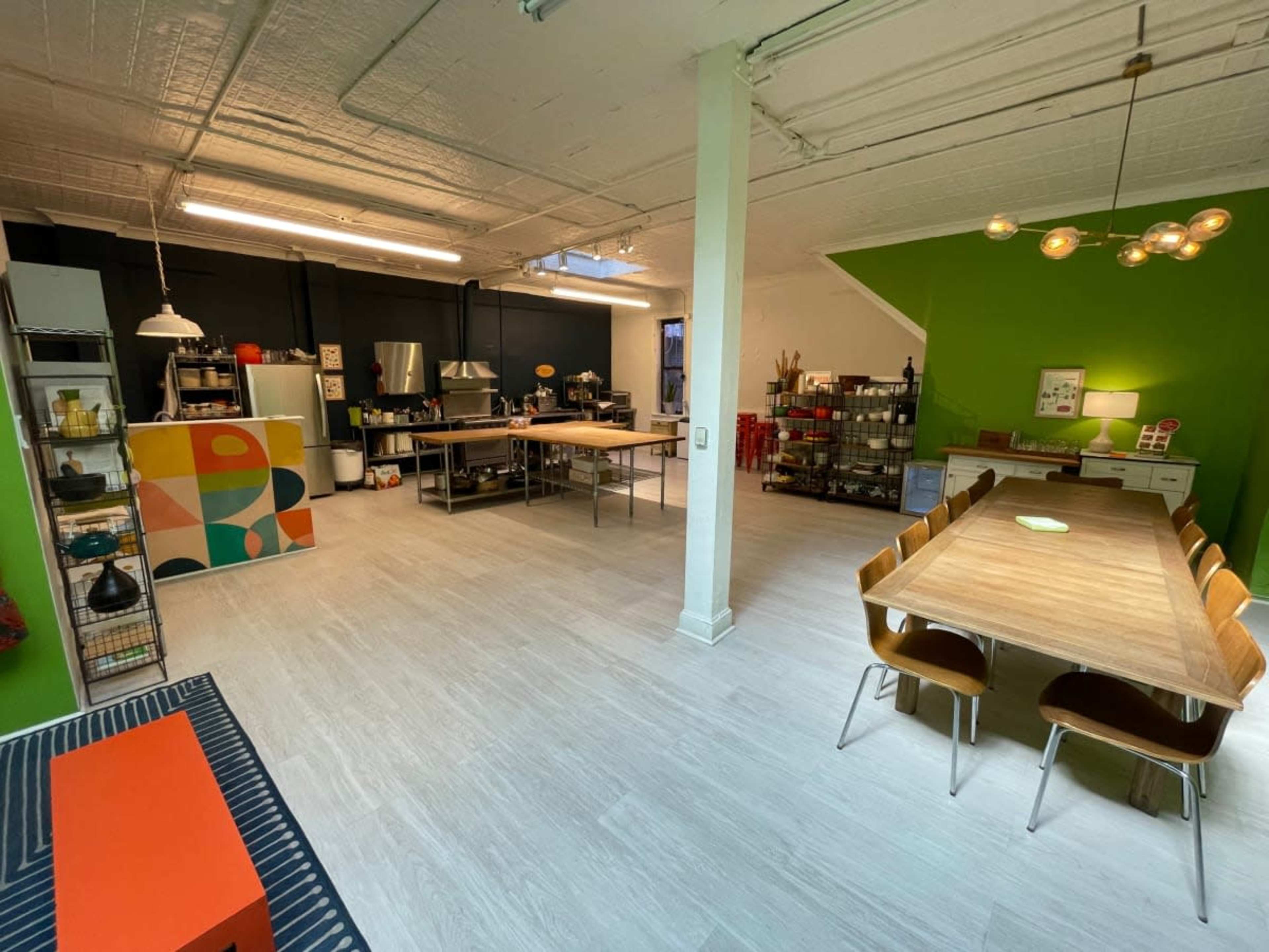 The image shows a spacious kitchen and dining area with a long wooden table, modern appliances, and colorful wall accents.