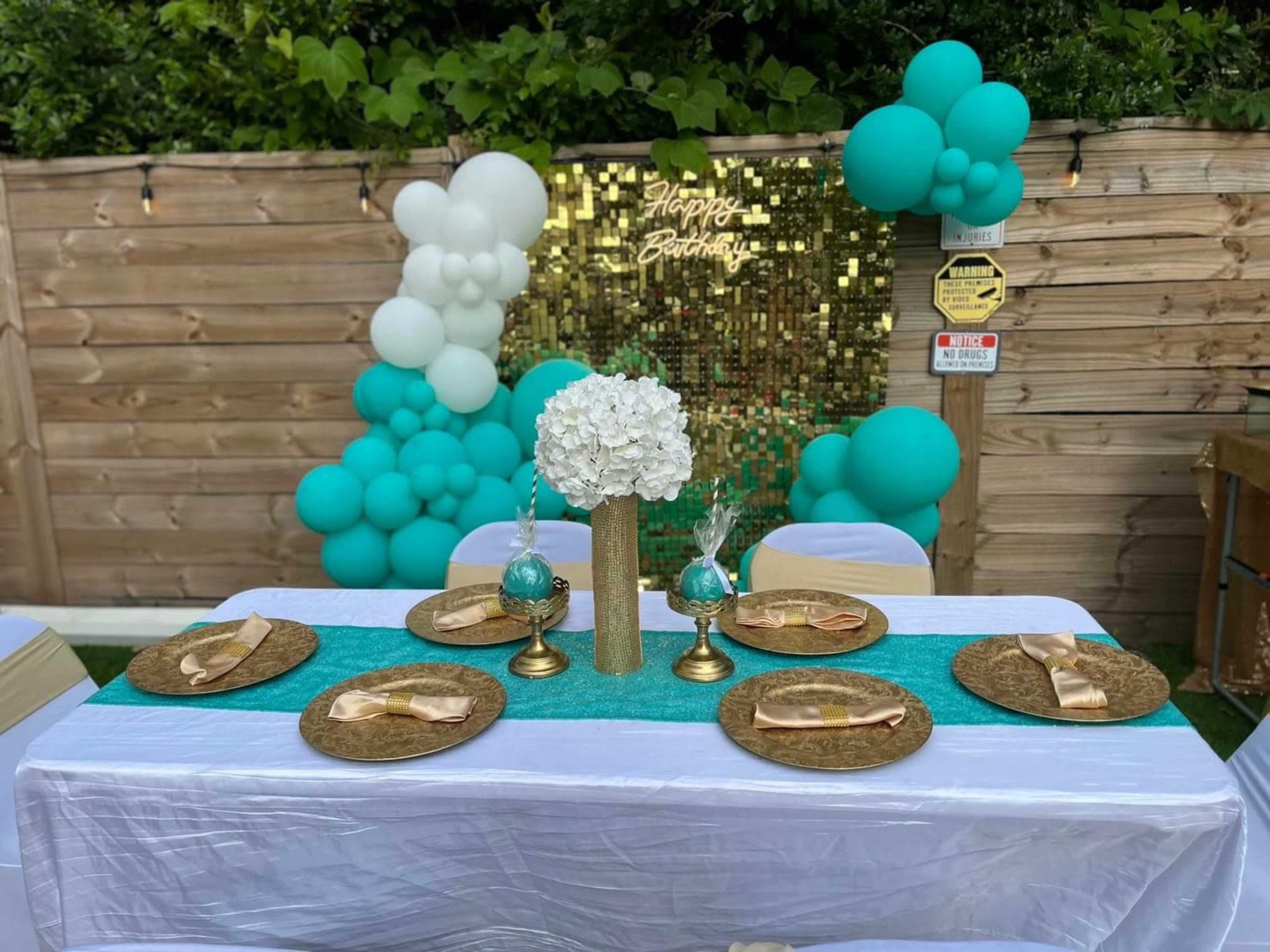 A birthday celebration setup features a decorated table with gold plates, a turquoise table runner, floral arrangements, and a backdrop of balloons.