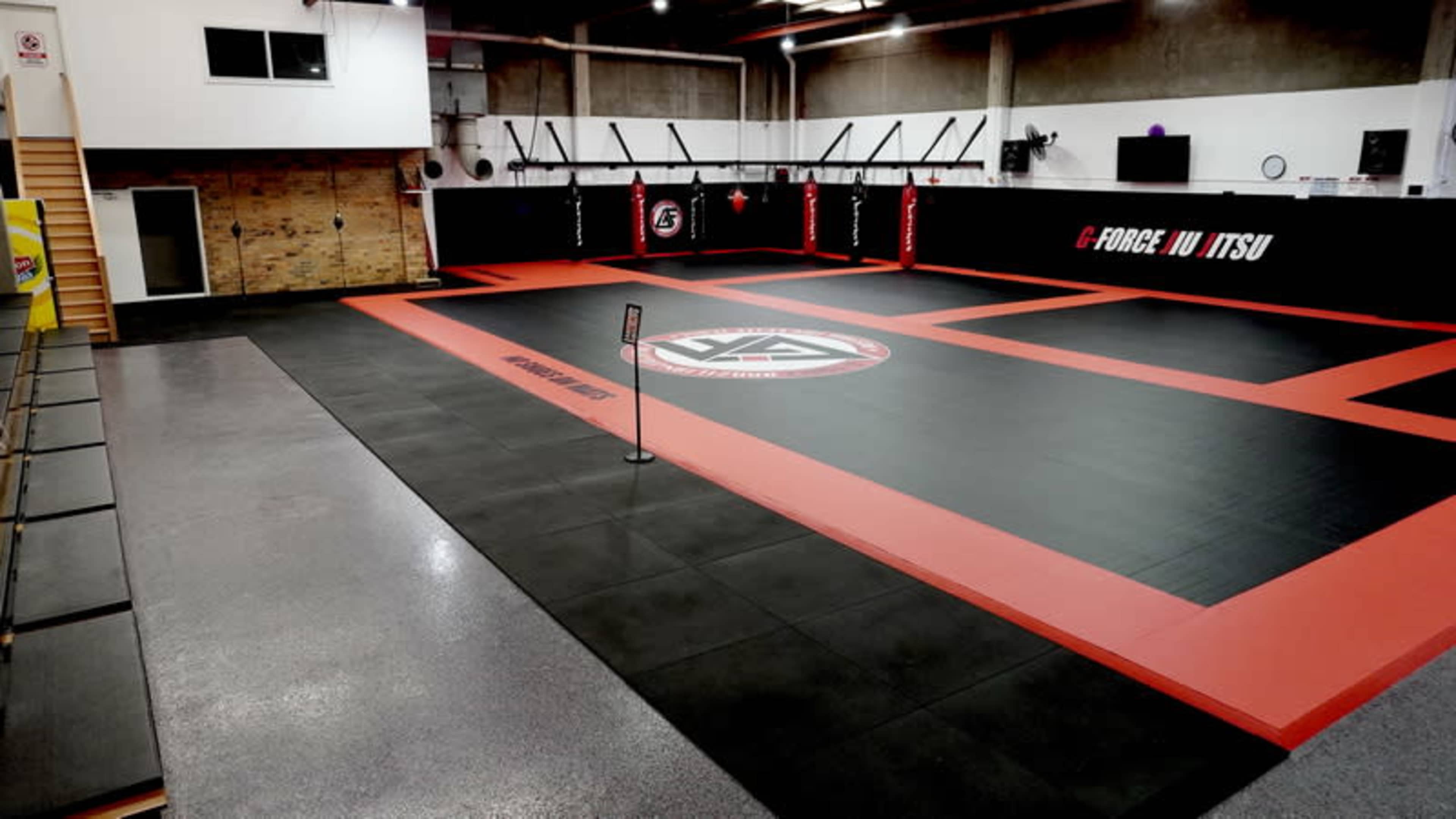 The image shows a spacious martial arts training gym with a black and red matted floor, surrounded by walls adorned with training equipment and branding.