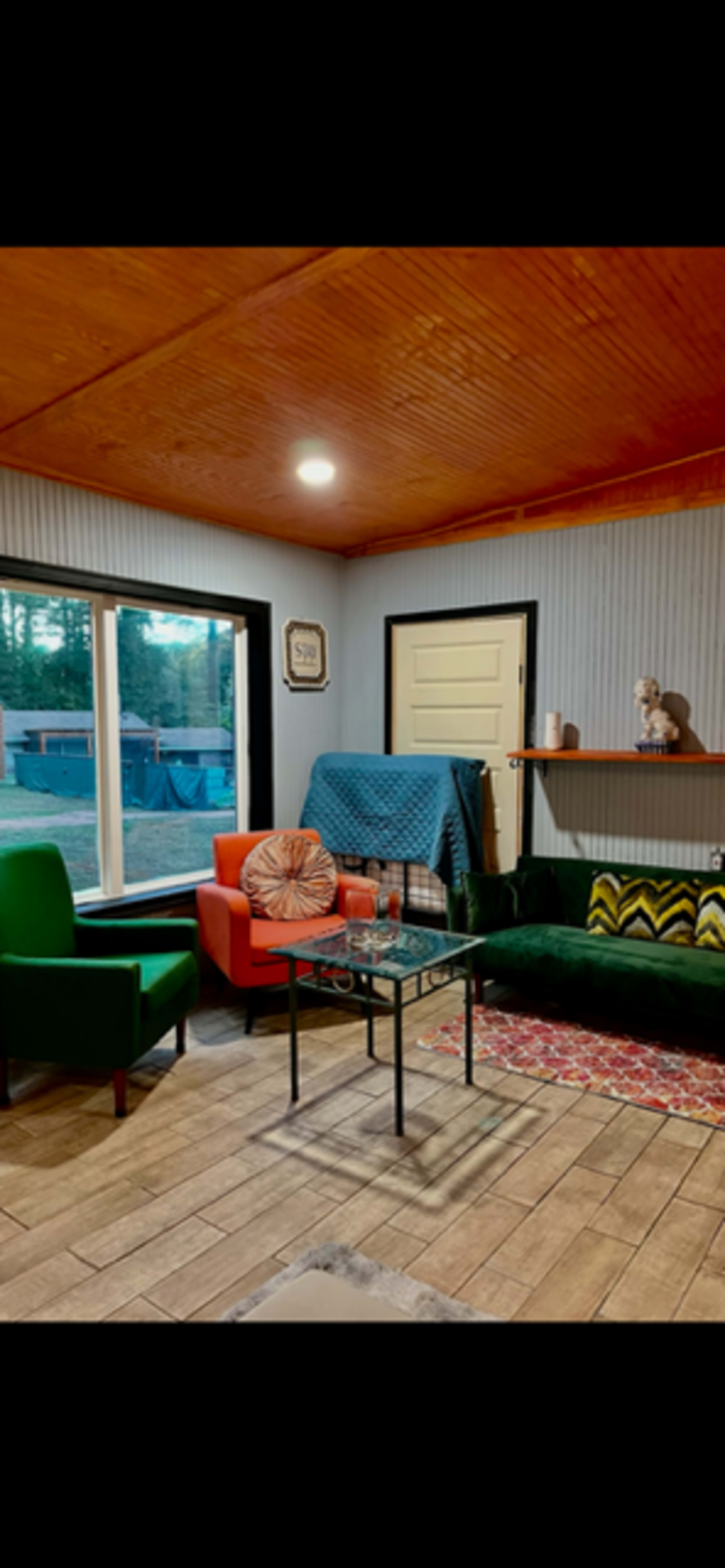 The image shows a cozy room with a green sofa, an orange chair, and a glass coffee table, featuring a window with a view of the outdoors and a shelf with decorative items.