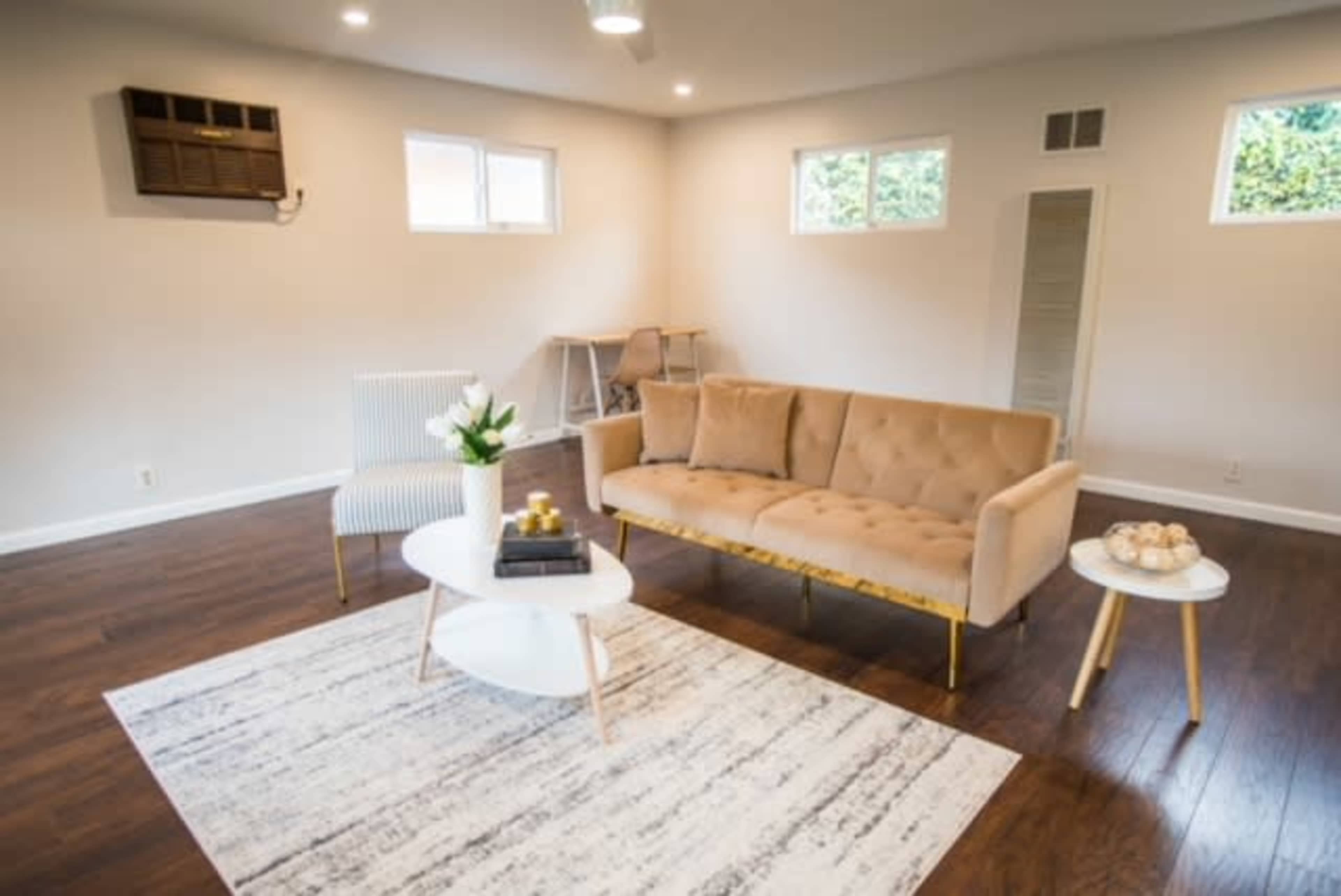 The image shows a modern living room with a beige tufted sofa, a small round coffee table, a patterned area rug, and large windows allowing natural light.