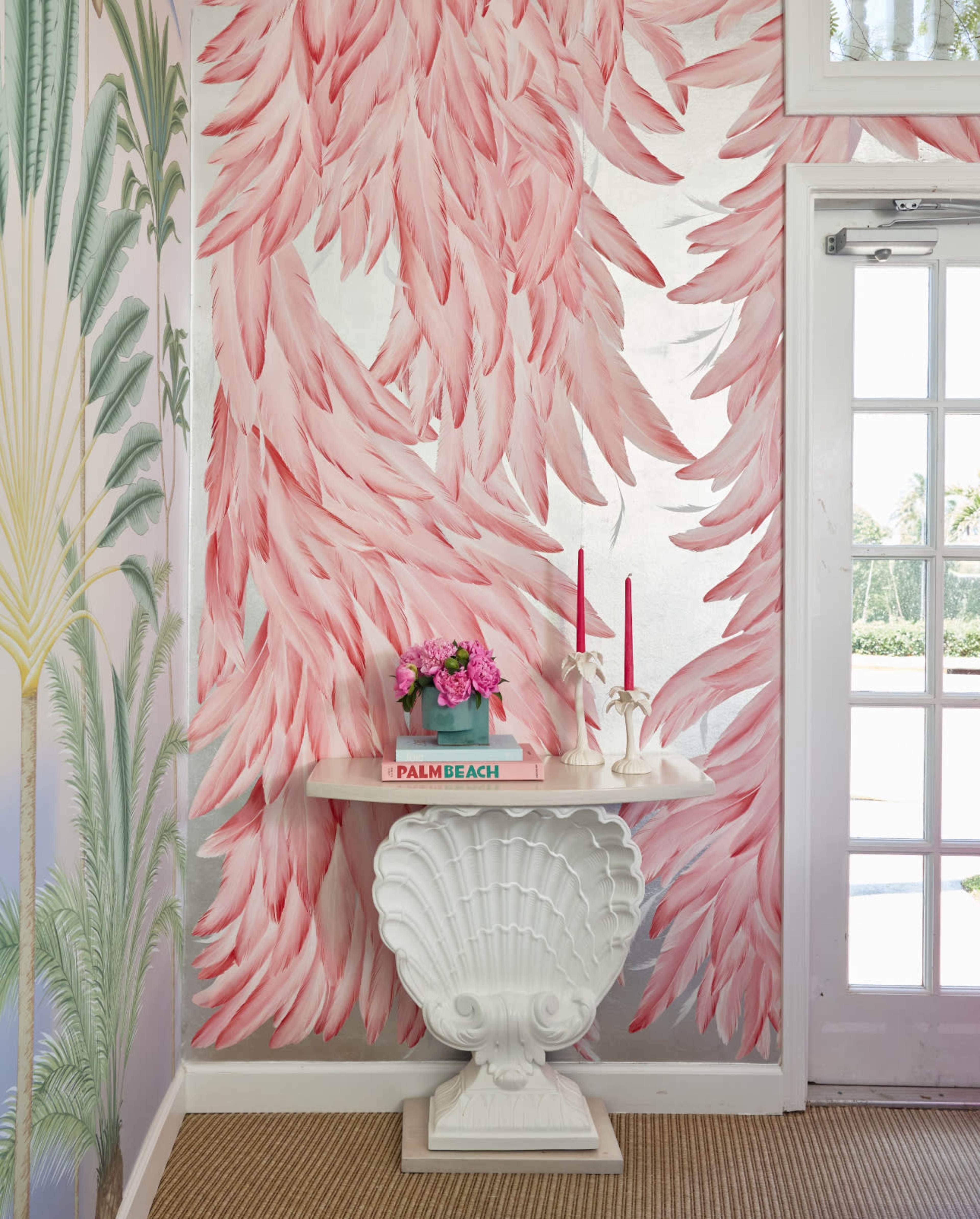 The image features a bright interior space with a pink feather-patterned wallpaper, a white shell-shaped table, and a floral arrangement on top, along with two pink candles.