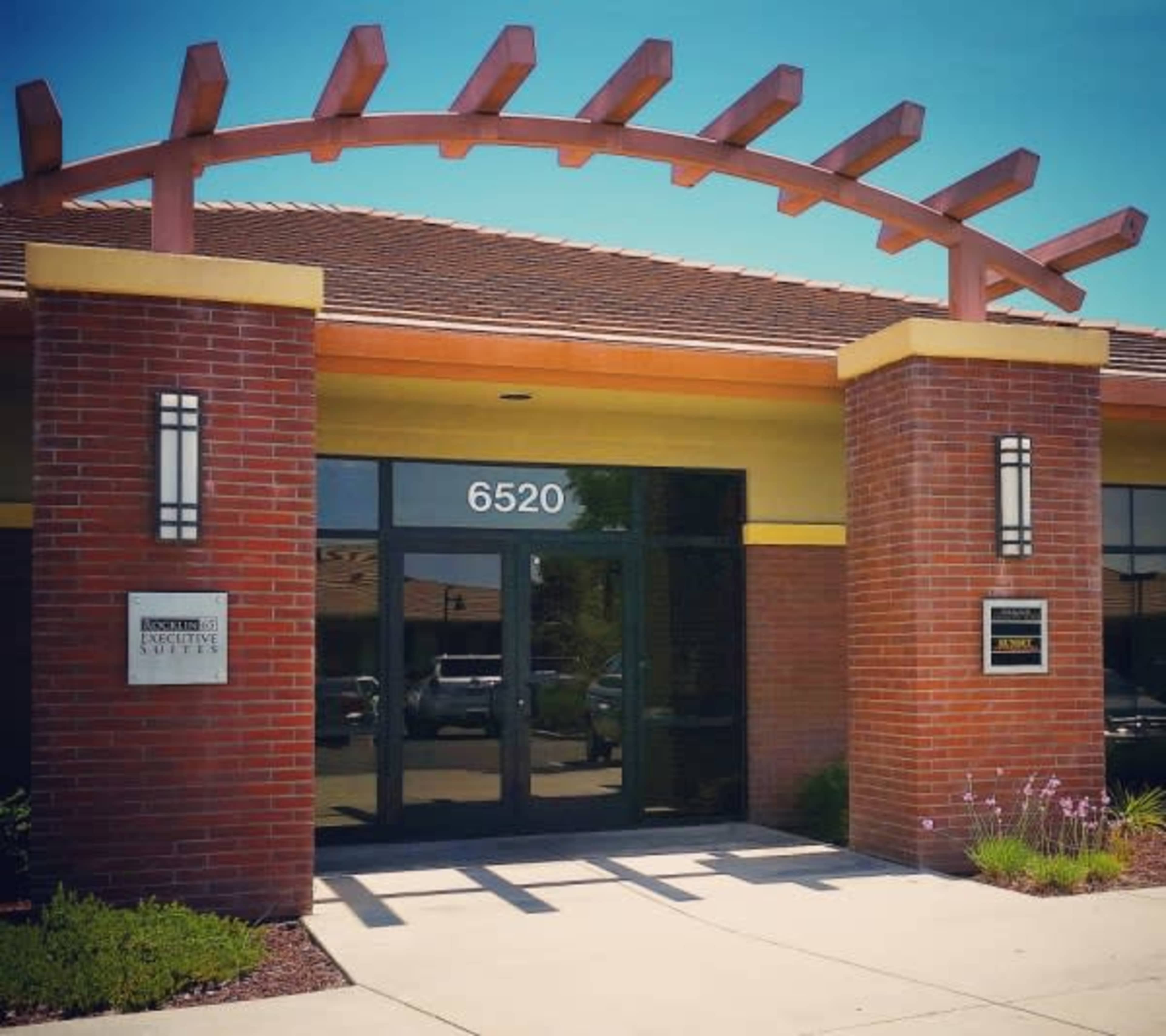 The entrance of a brick building with a decorative arch, featuring large glass doors and signs indicating room numbers and the name of the institution.