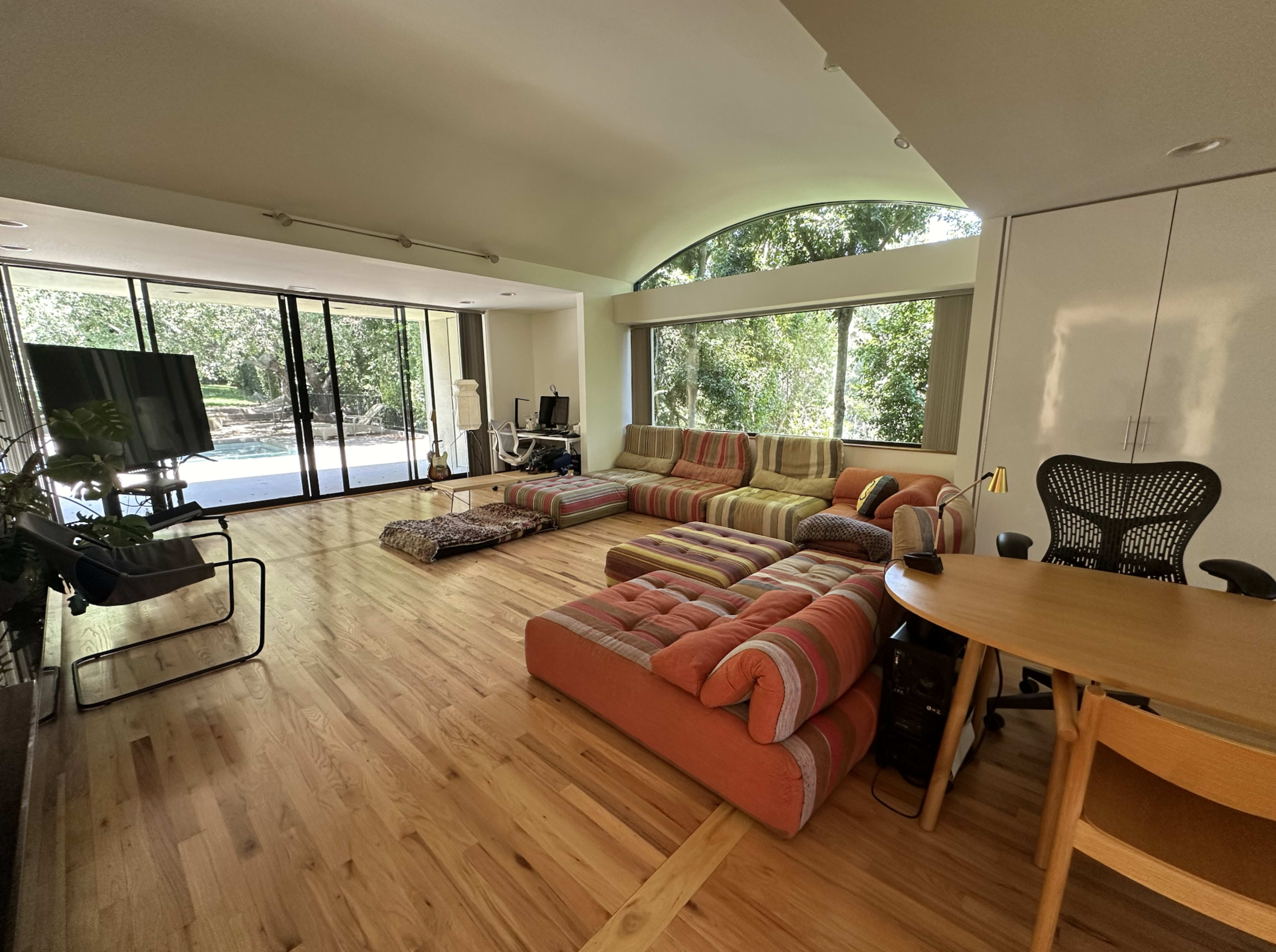The image shows a spacious living room with a large striped sectional sofa, a wooden table, and large windows overlooking a green outdoor area.
