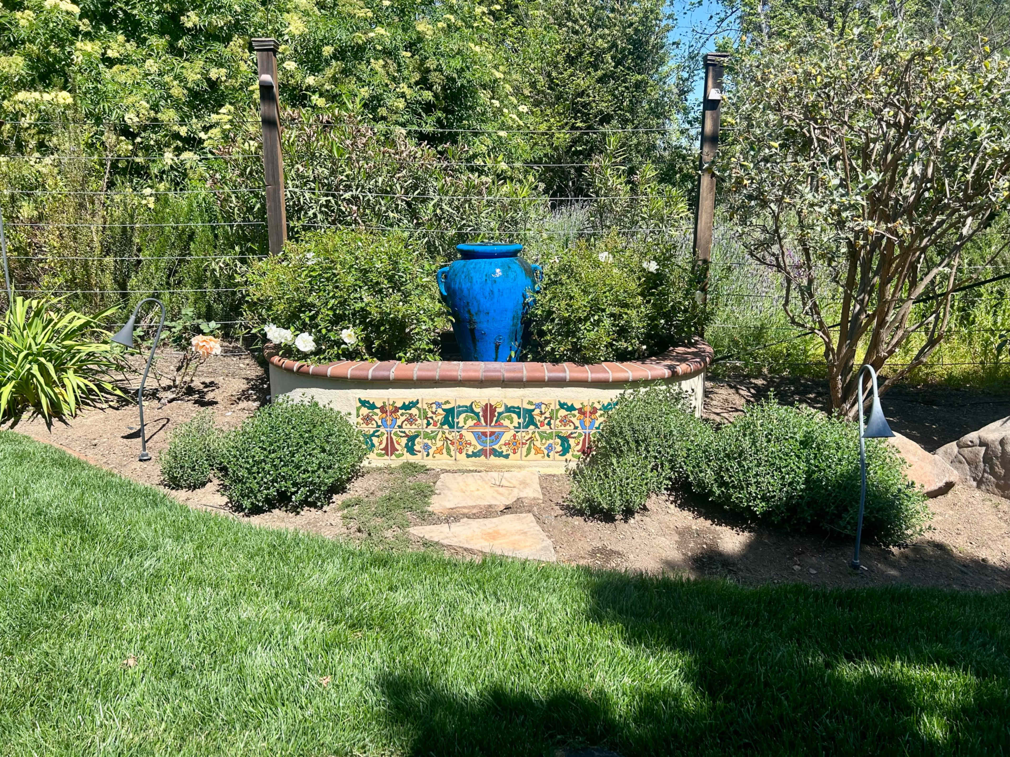 A blue ceramic pot sits atop a garden wall decorated with colorful tiles, surrounded by lush greenery and a stone pathway.