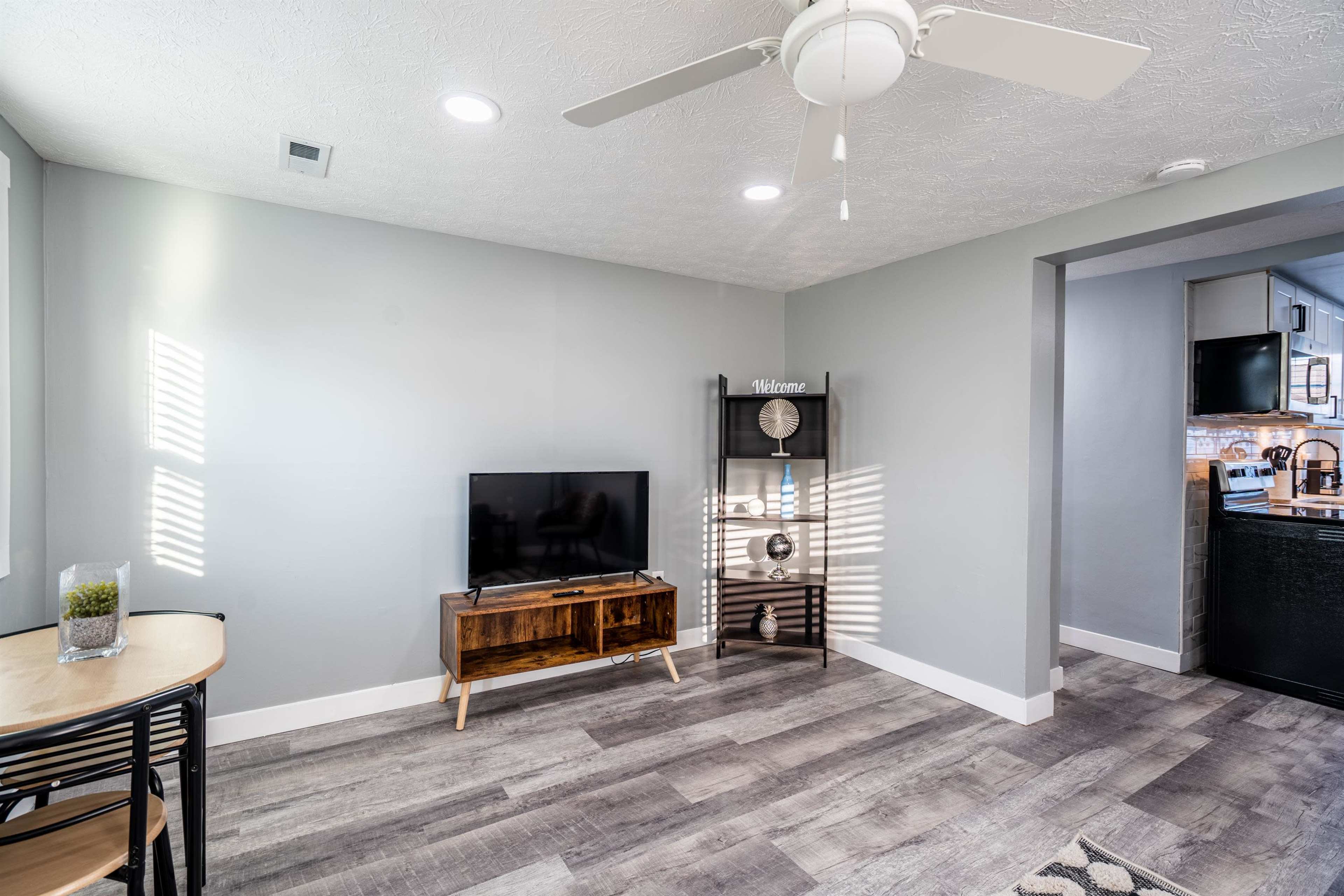 A living space features a television, a small dining table, and a wall-mounted shelf in front of a light gray backdrop with natural light streaming in through nearby windows.