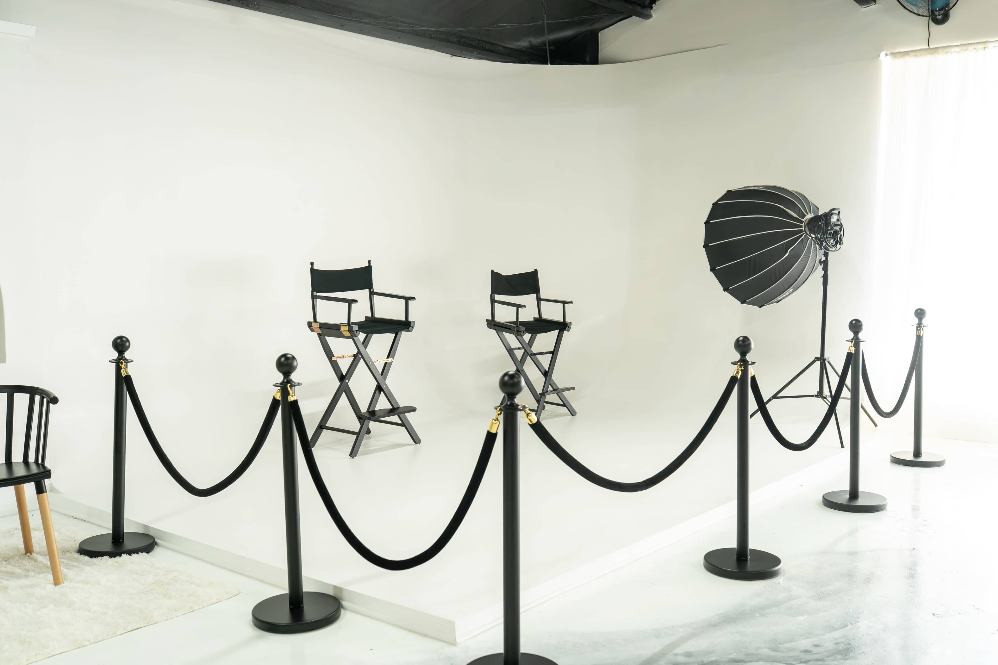 The image shows a minimalist photo studio setup with two director's chairs, a large softbox light, and a rope barrier surrounding the area.