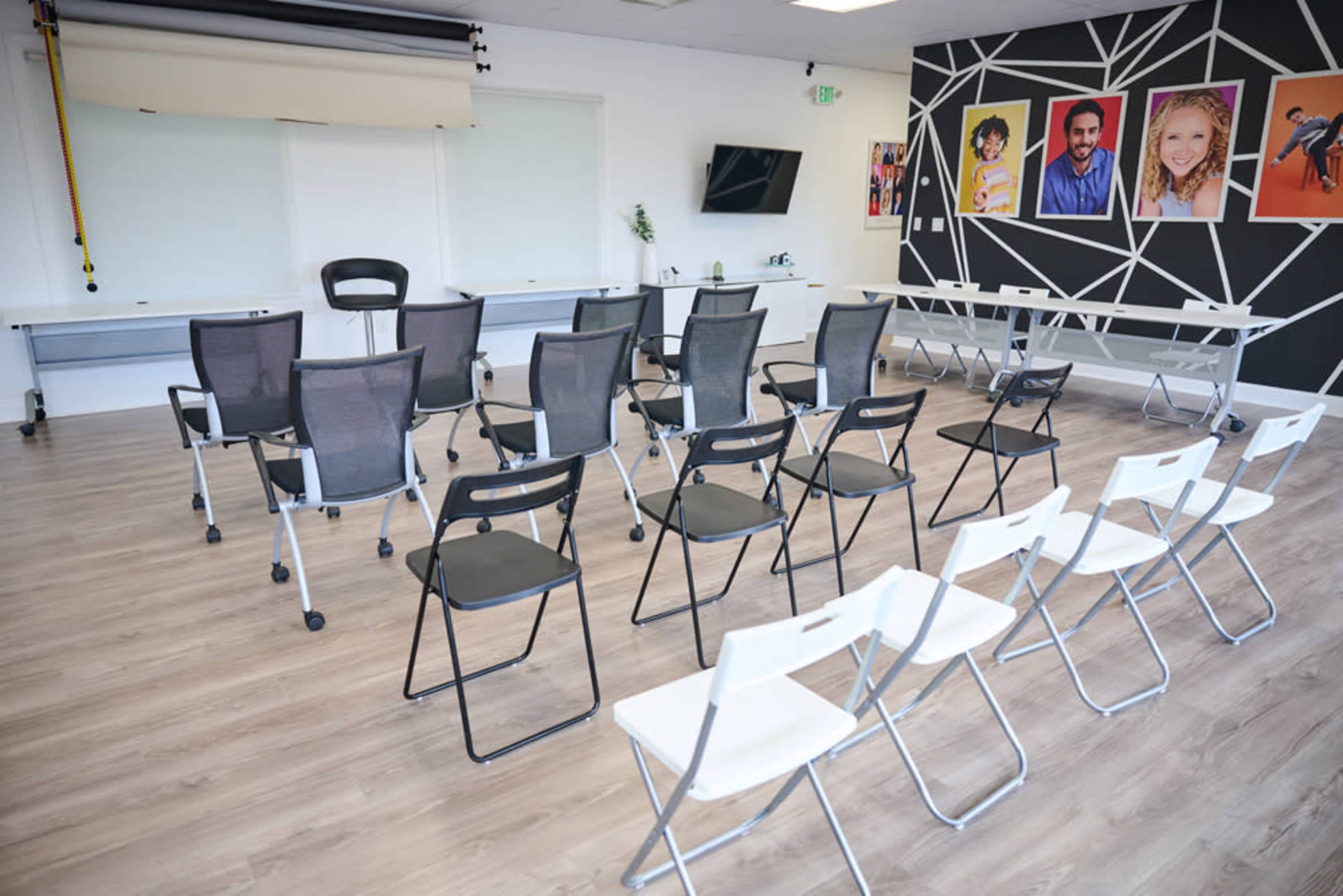 The image shows a brightly lit meeting room with rows of chairs arranged in an open space, accompanied by a wall featuring colorful portraits.