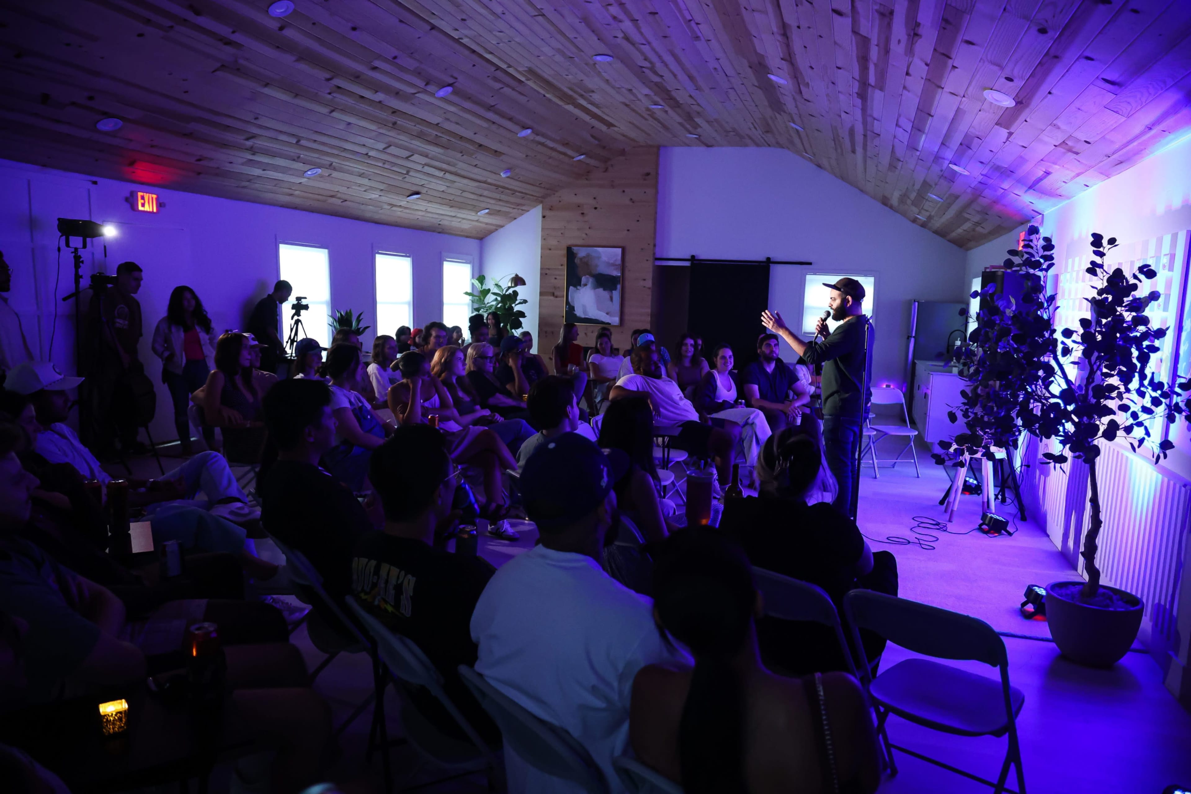 A performer stands on stage in a softly lit room filled with an audience seated in chairs, attentively watching the show.