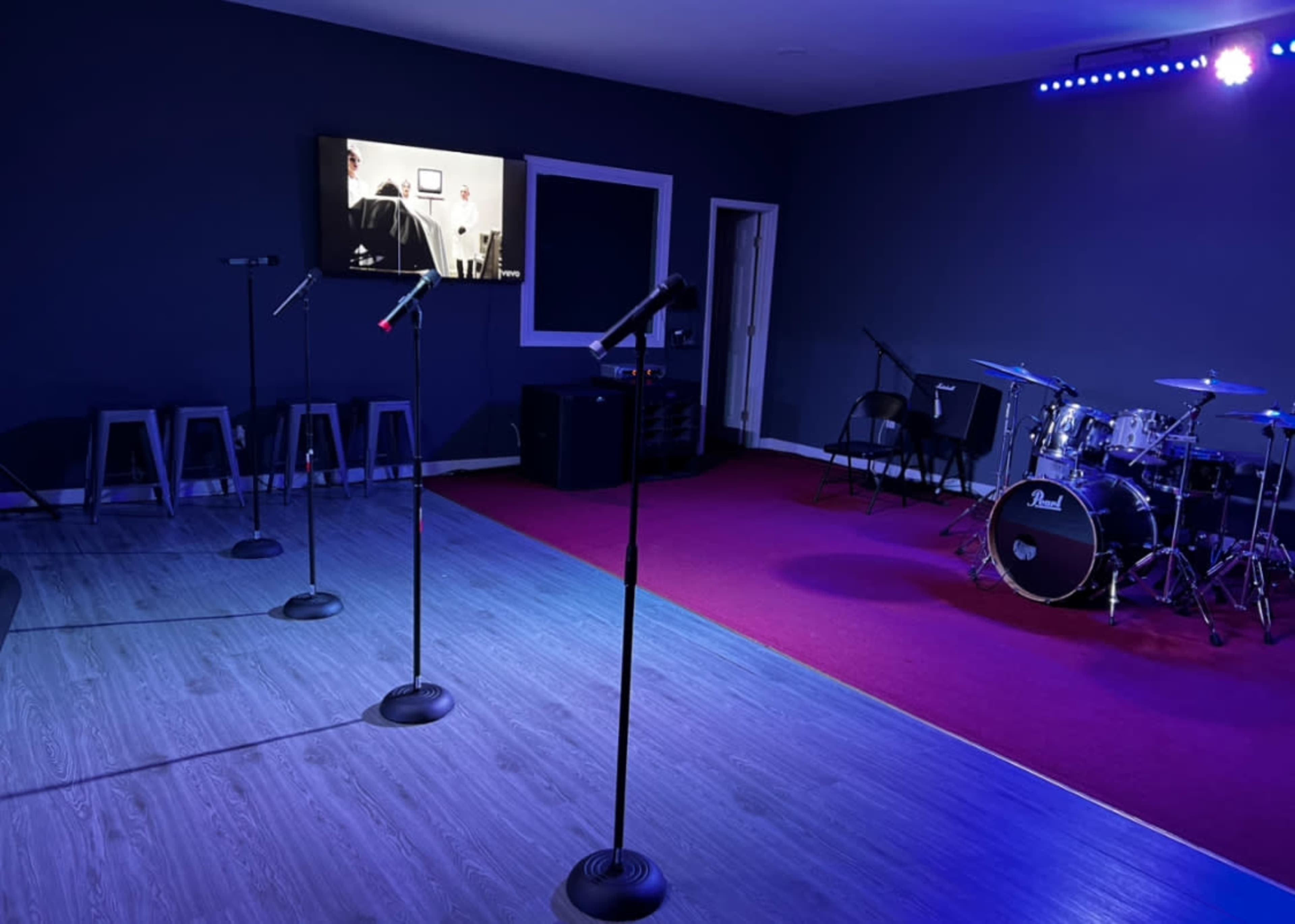 The image shows a performance space equipped with several microphone stands, a television screen, and a small drum set.