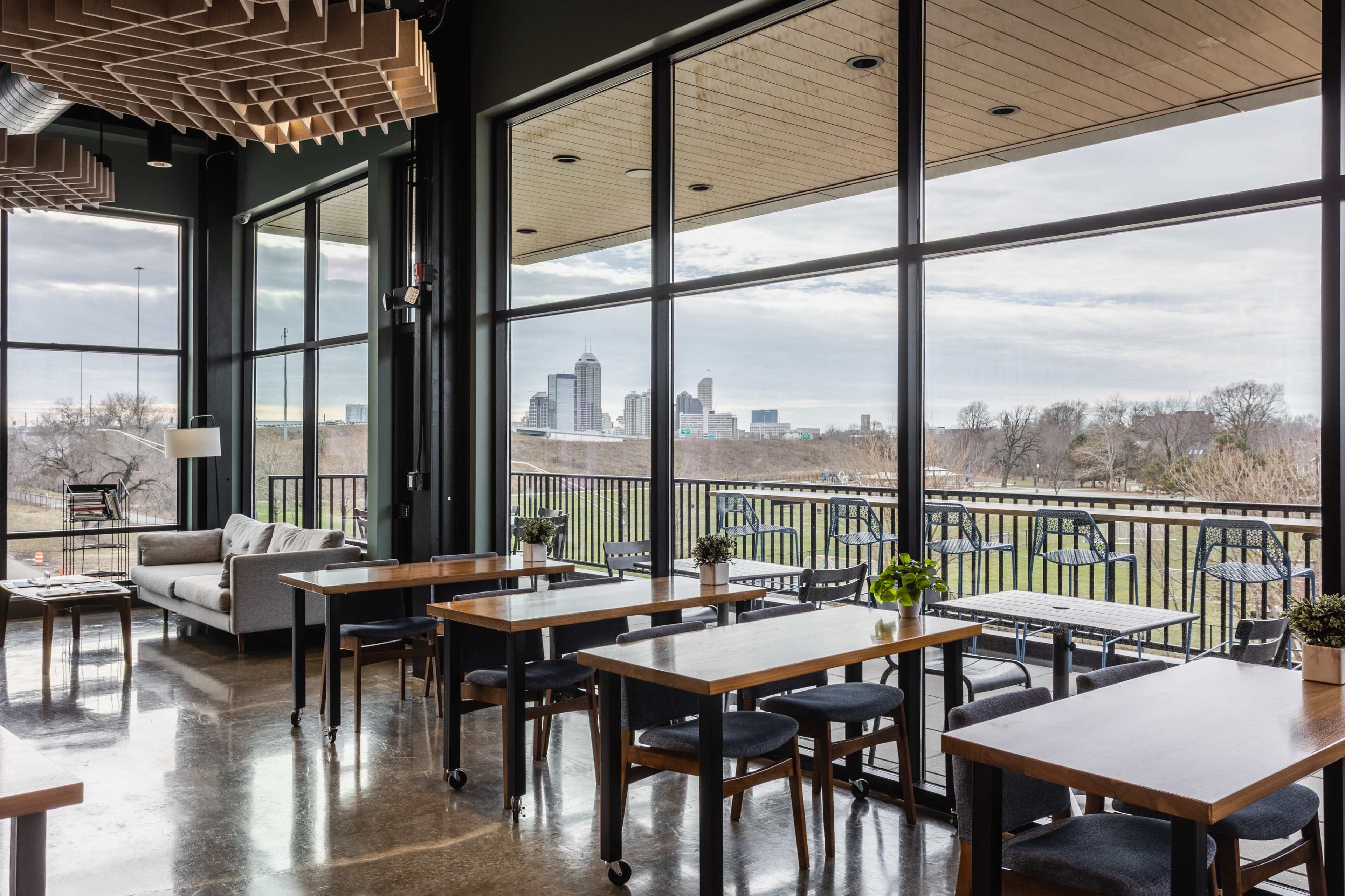 The image shows a modern café with large windows offering a view of a city skyline, furnished with wooden tables and chairs.