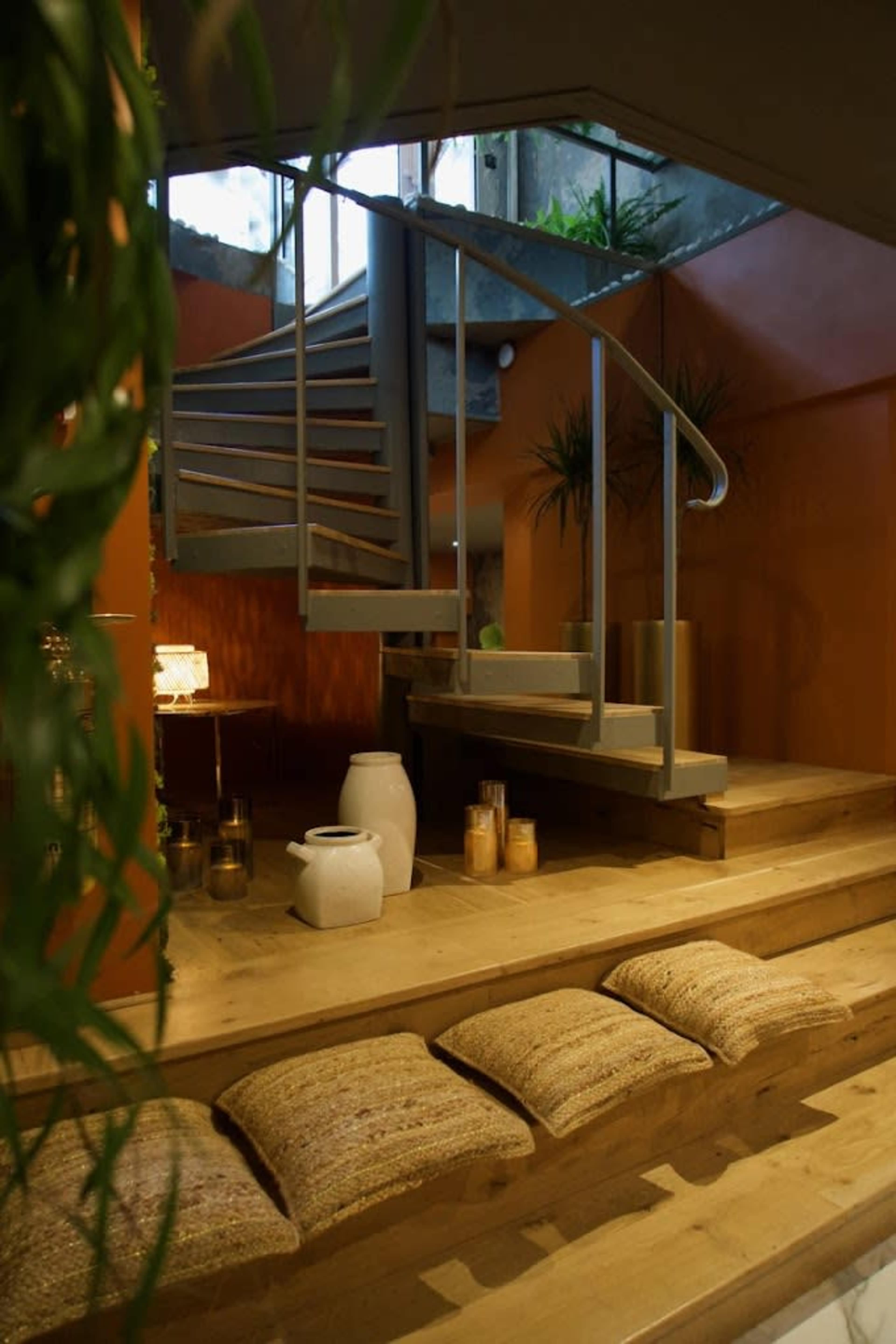 The image shows a modern interior with a spiral staircase leading up, surrounded by plants and decorated with wooden elements and soft seating.