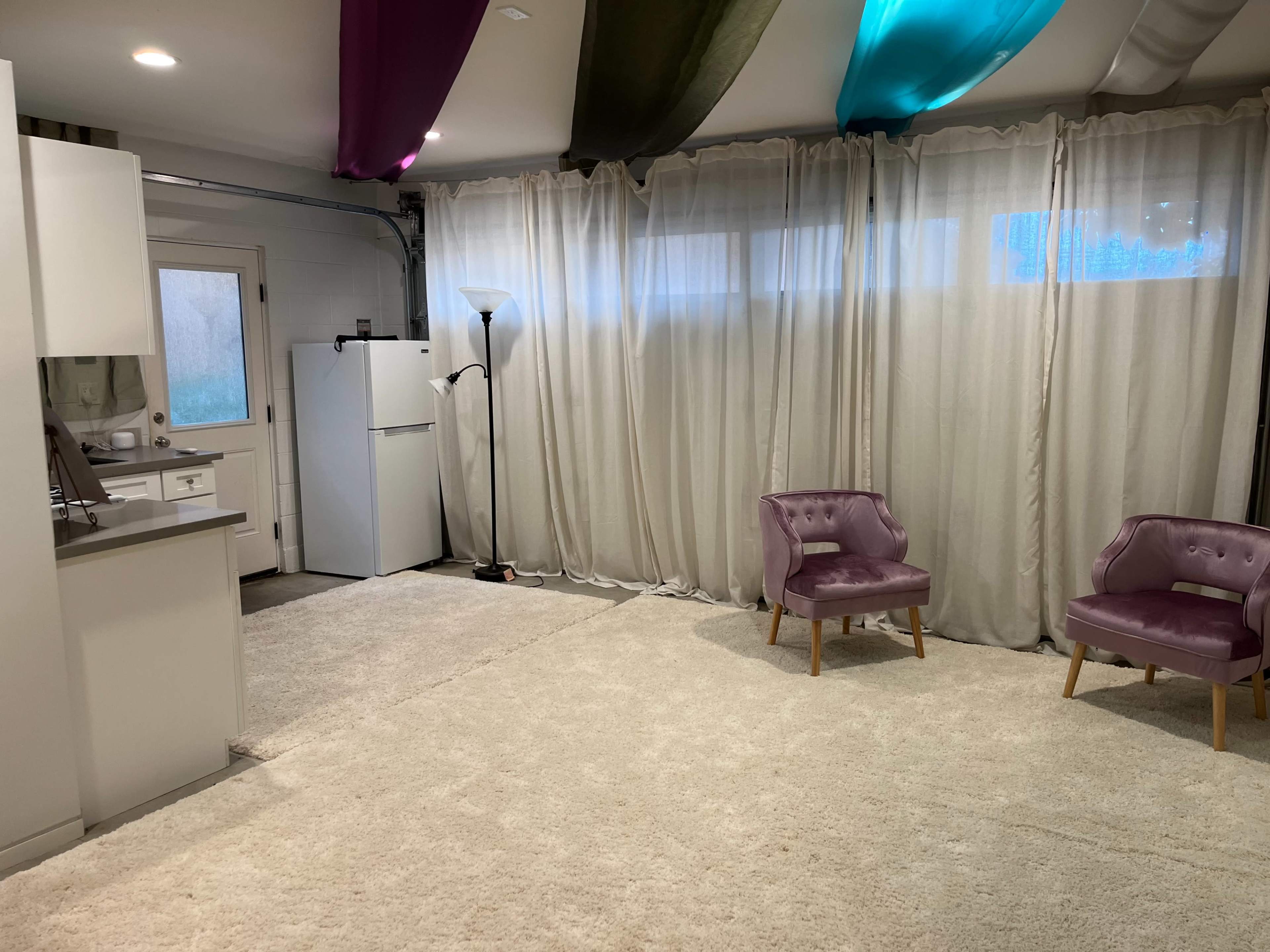 The image shows an indoor space with two purple chairs, a white refrigerator, and sheer curtains draping the walls over a light-colored carpet.