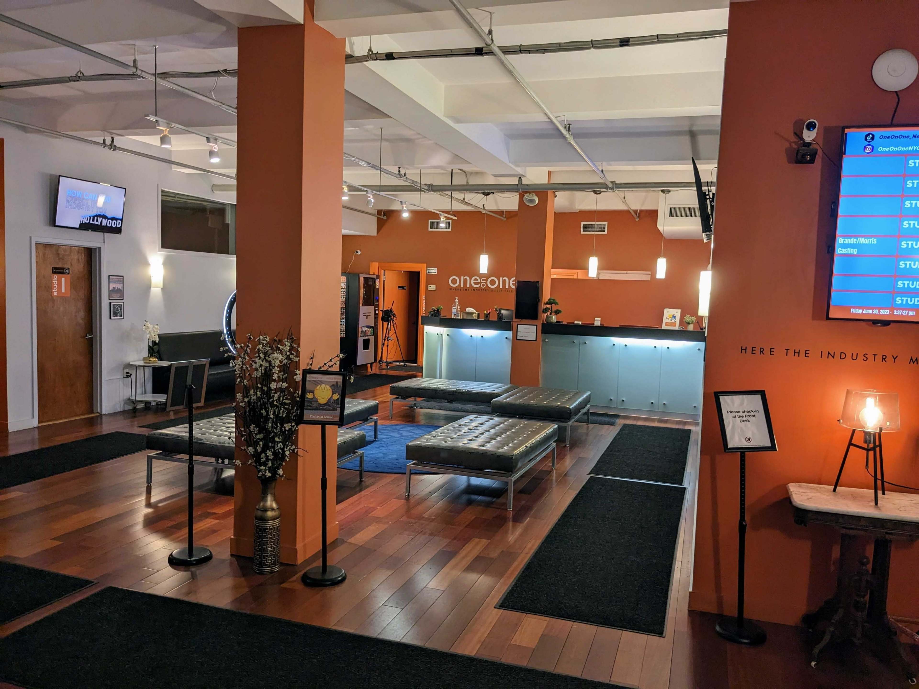 The image depicts a modern reception area with polished wooden floors, an orange accent wall, and several seating areas featuring silver benches.