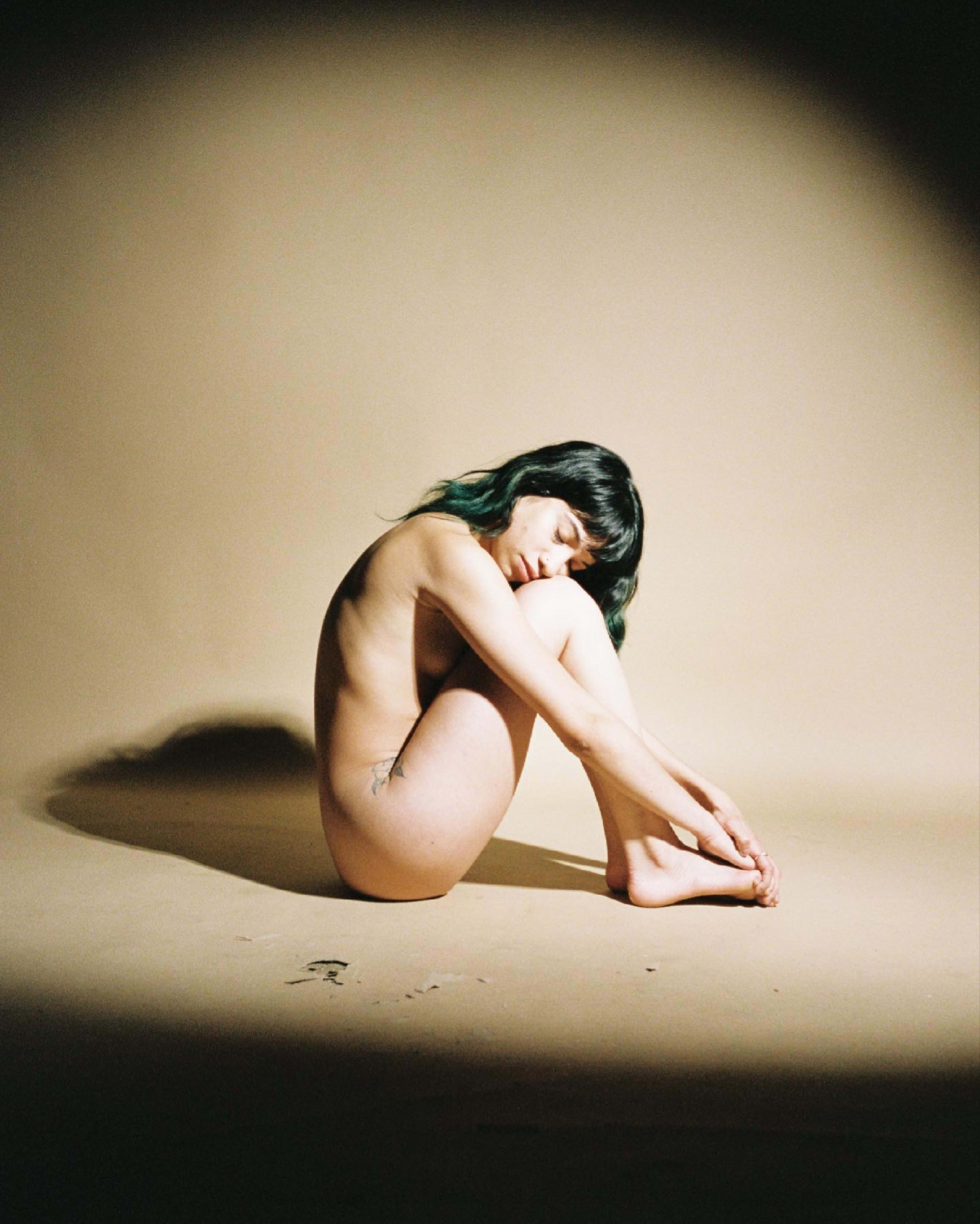 A minimalist boudoir photoshoot featuring a nude woman sitting cross-legged on the ground.