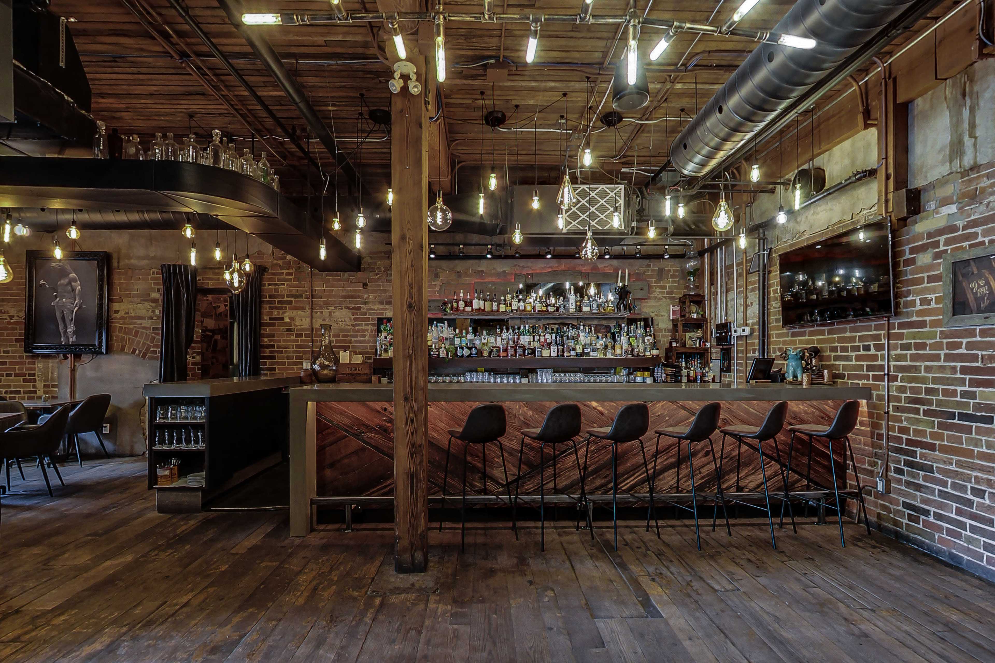 The image shows a rustic bar interior with exposed brick walls, wooden beams, and a long bar counter lined with modern stools.