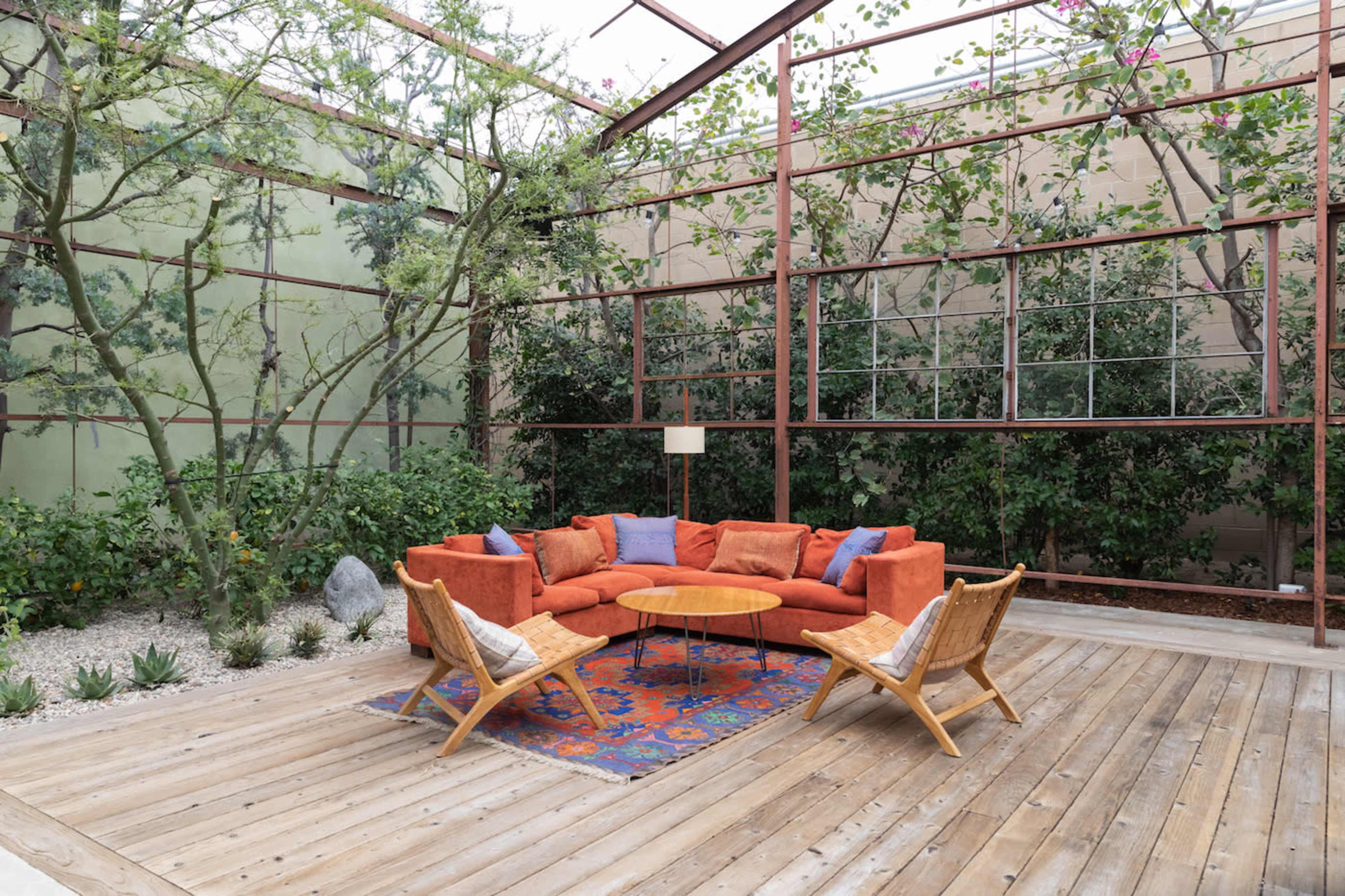 A modern outdoor seating area features an orange sectional couch, a wooden coffee table, and two wicker chairs, surrounded by greenery and large windows.