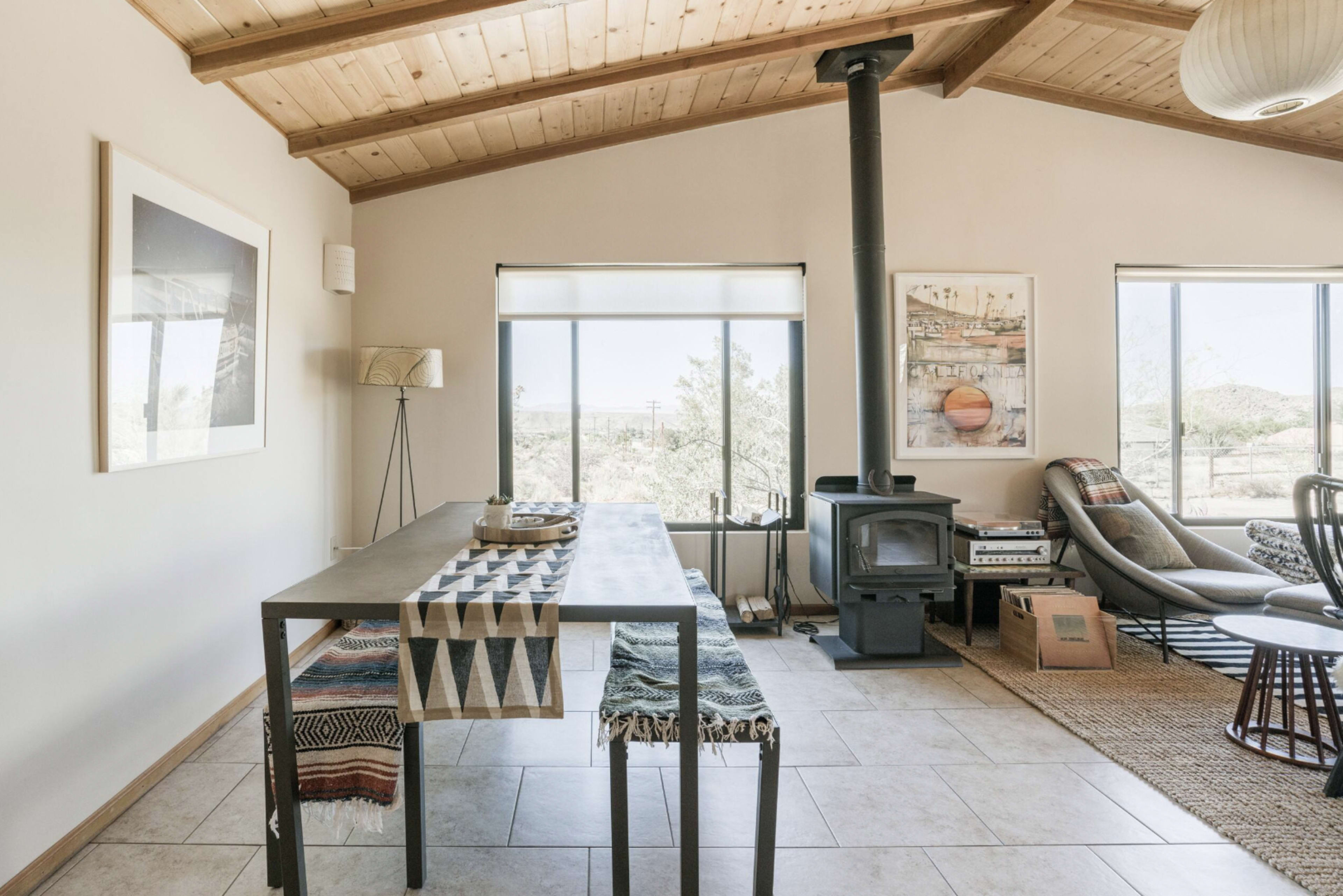 The image shows a simple dining area with a metal table and a wood-burning stove in a room featuring large windows and a wooden ceiling.